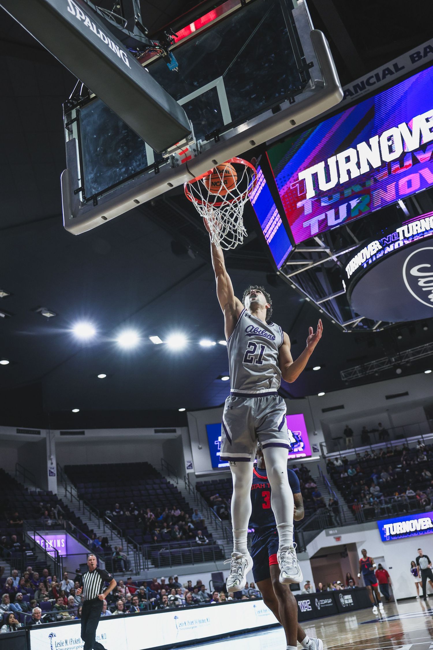 College basketball player #21 drives to the basket for a layup as scoreboard displays TURNOVER in purple.