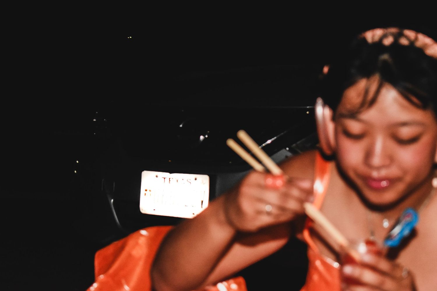 A young girl in an orange outfit holds chopsticks in a dark setting with a blurred white card visible.
