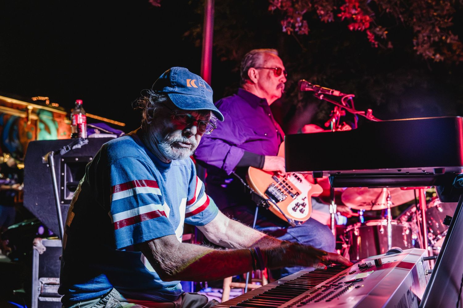 Two musicians performing outdoors at night, one playing keyboard and one playing guitar on stage.
