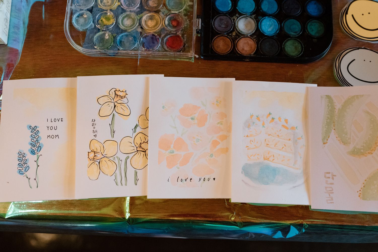 Handmade watercolor greeting cards with floral designs and love messages displayed on a table near a paint palette.