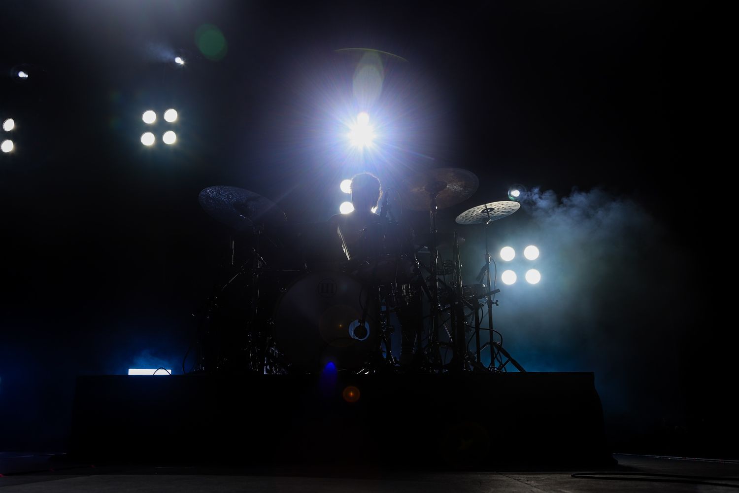 Empty drum kit on dark stage illuminated by bright white spotlights and blue smoke at a concert venue.