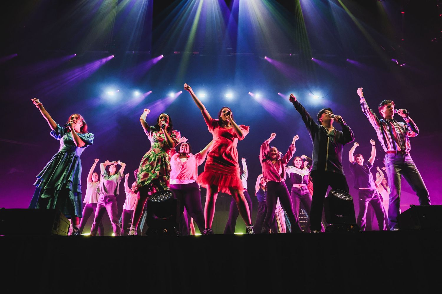Large cast of performers on stage with arms raised under colorful concert stage lighting.