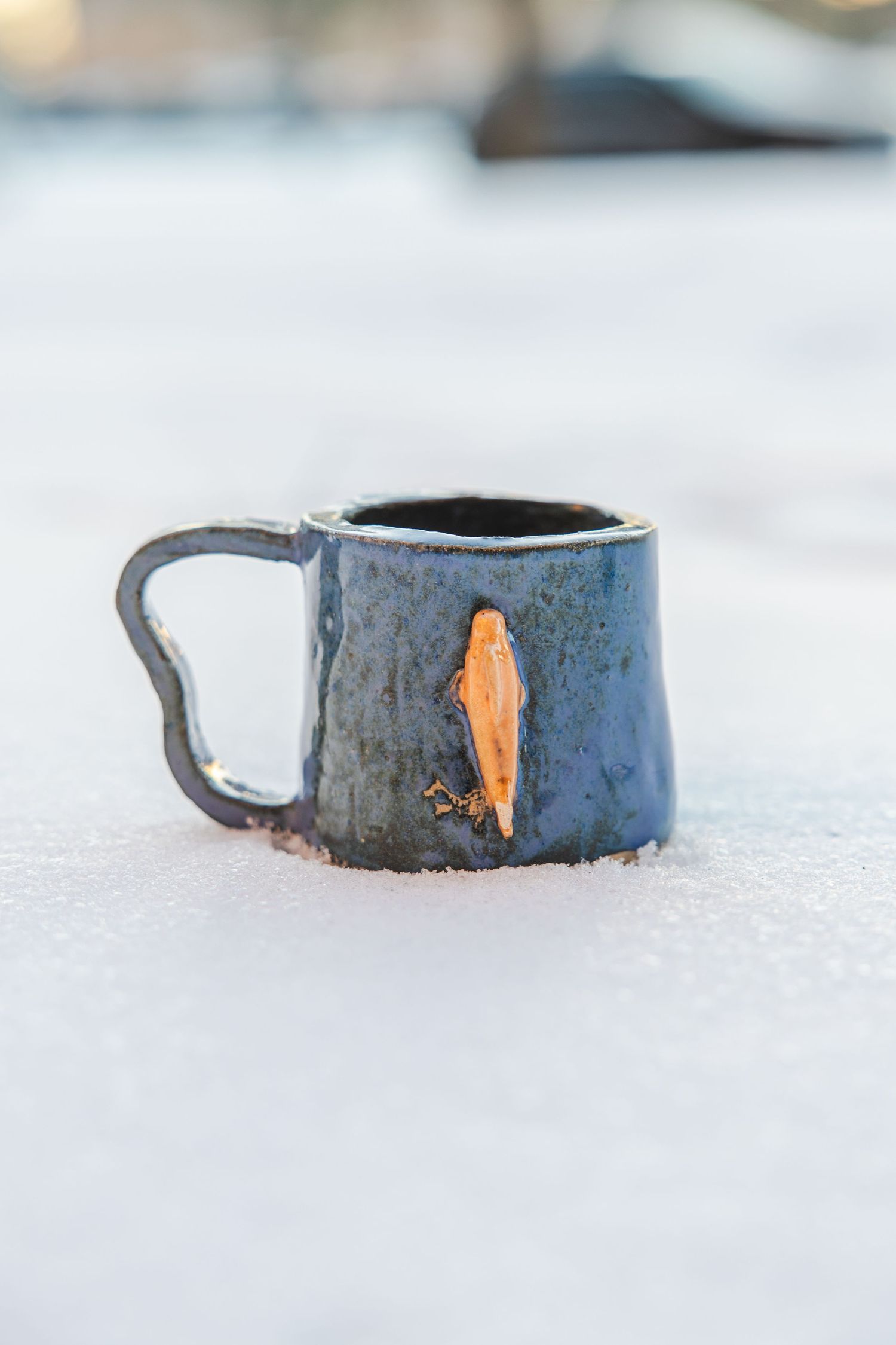 Dark blue glazed ceramic mug with orange leaf detail sitting in white snow.