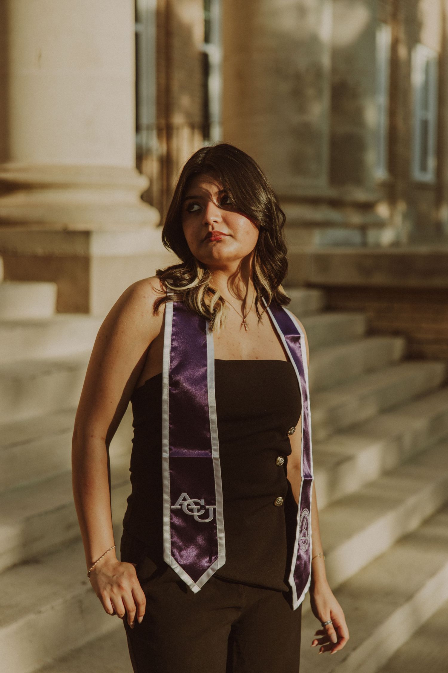 Young woman wearing purple ACU graduation stole standing on steps in golden sunlight.
