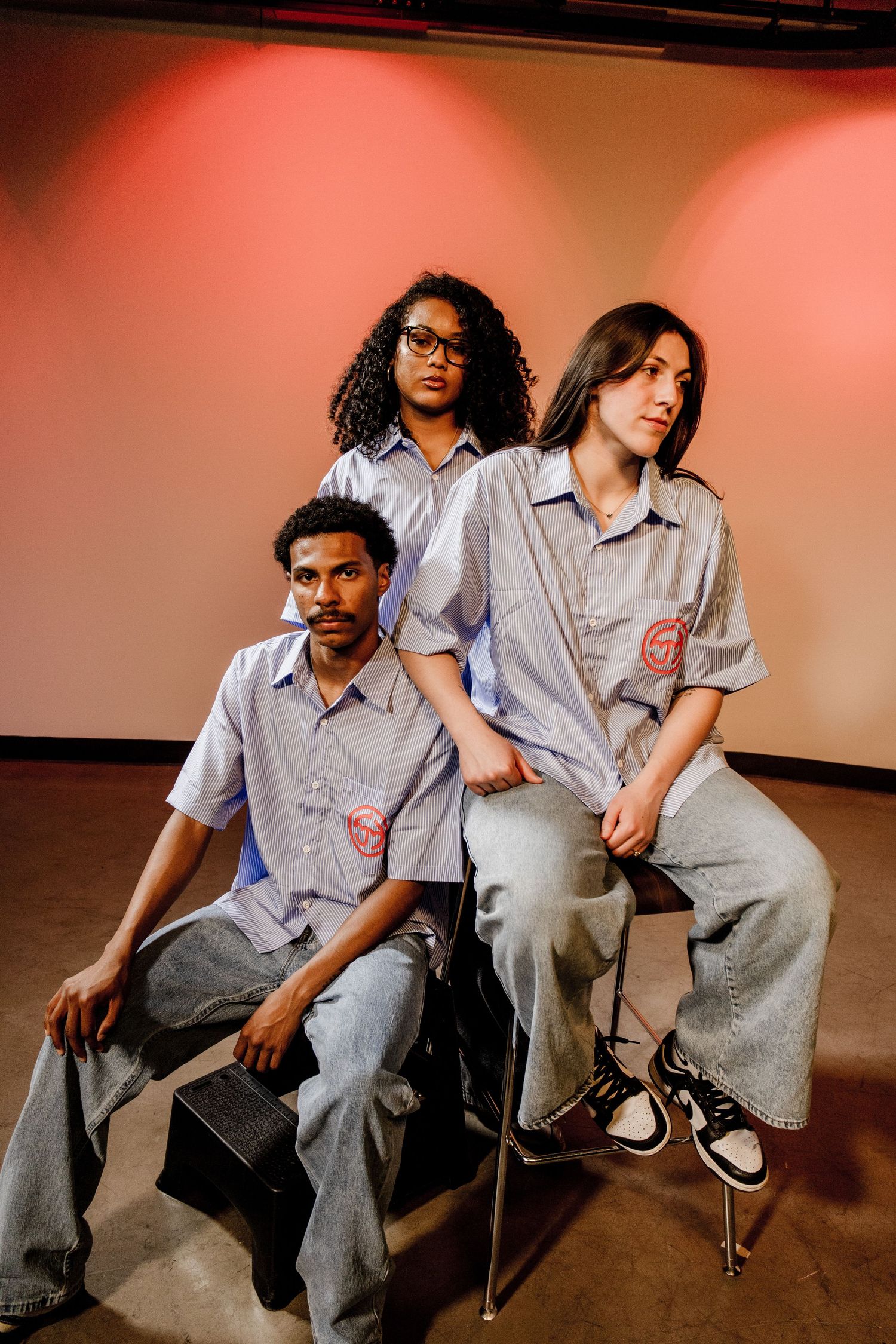 Three models pose in matching blue striped shirts with red logos in warmly lit studio.
