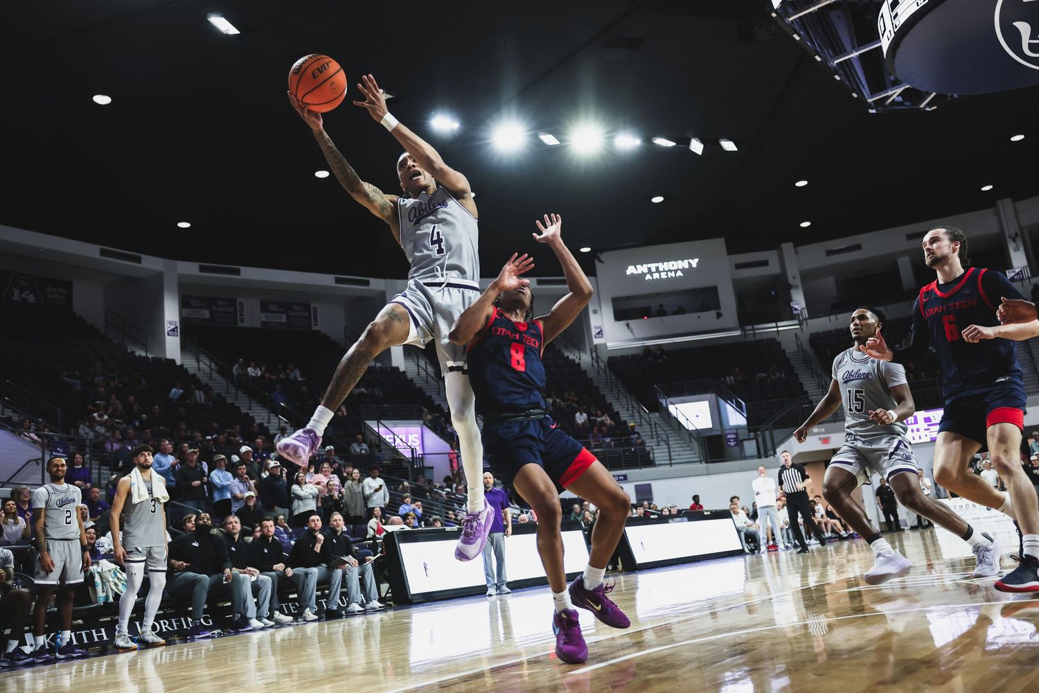 A college basketball player in white leaps to shoot over a red-uniformed defender in a packed indoor arena.