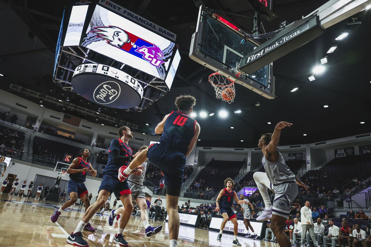College basketball game action shot showing players near the hoop under bright arena lights with scoreboard visible.
