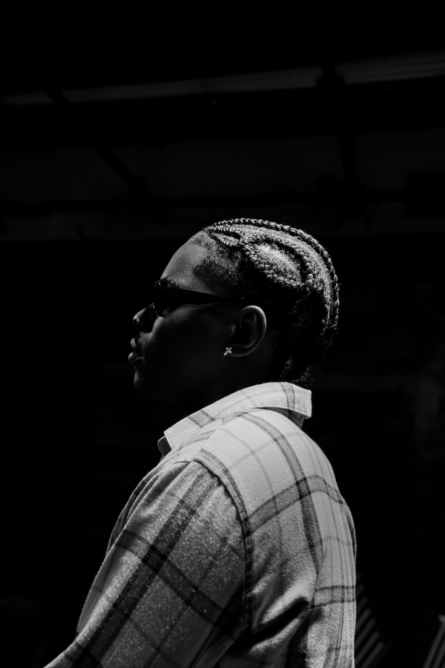 Black and white profile portrait of a person with braided hair wearing a plaid shirt in dark lighting.