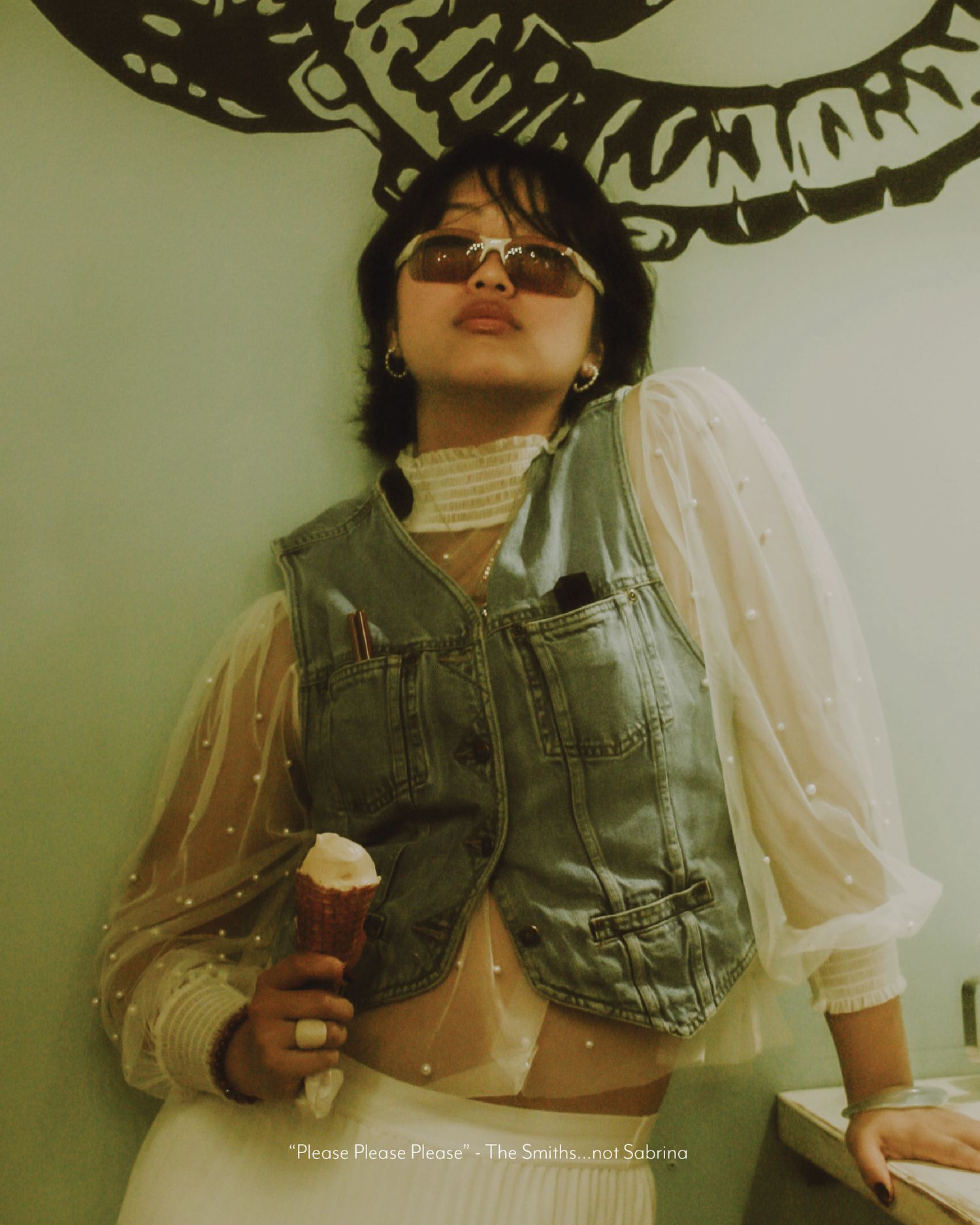 Young woman in denim vest and sheer blouse holding ice cream, wearing sunglasses indoors.