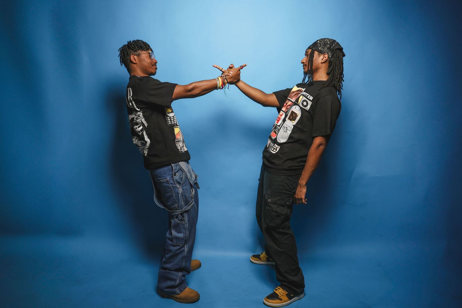 Two men in matching black graphic tees and baggy jeans doing a handshake against blue backdrop.