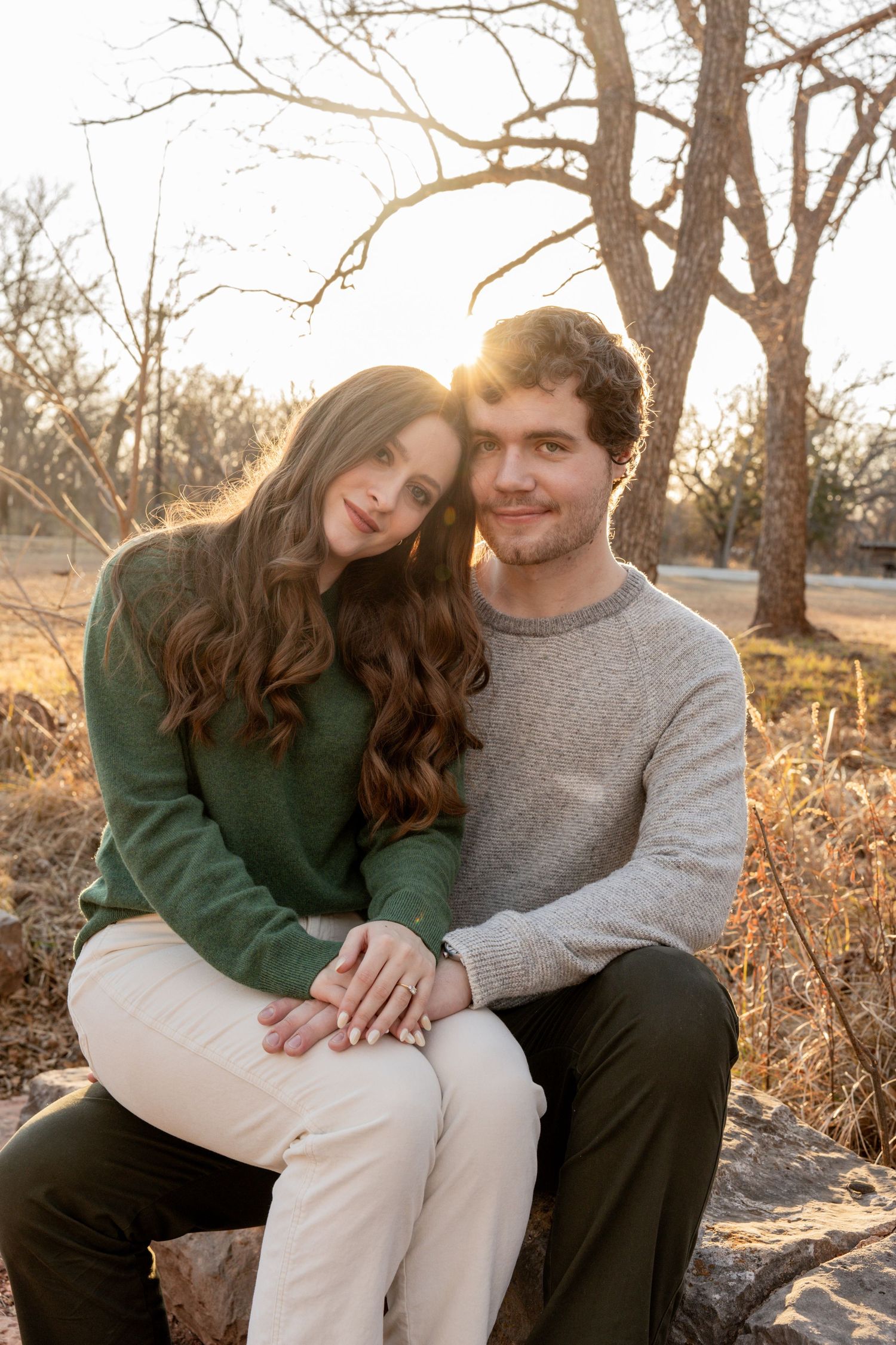Couple sitting on a rock at golden hour, woman in green sweater leaning on man in gray sweater outdoors.