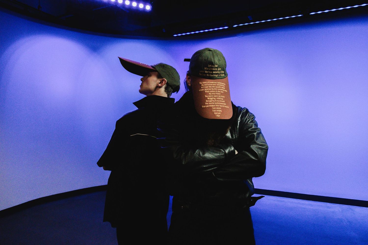 Two people in black coats and caps pose in blue-lit room, one wearing text-covered mask.