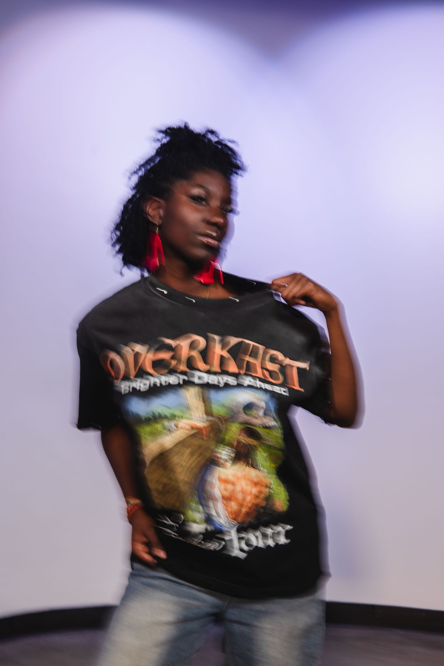 Young woman wearing a black Overkast Brighter Days Ahead tour graphic t-shirt with red earrings and jeans.