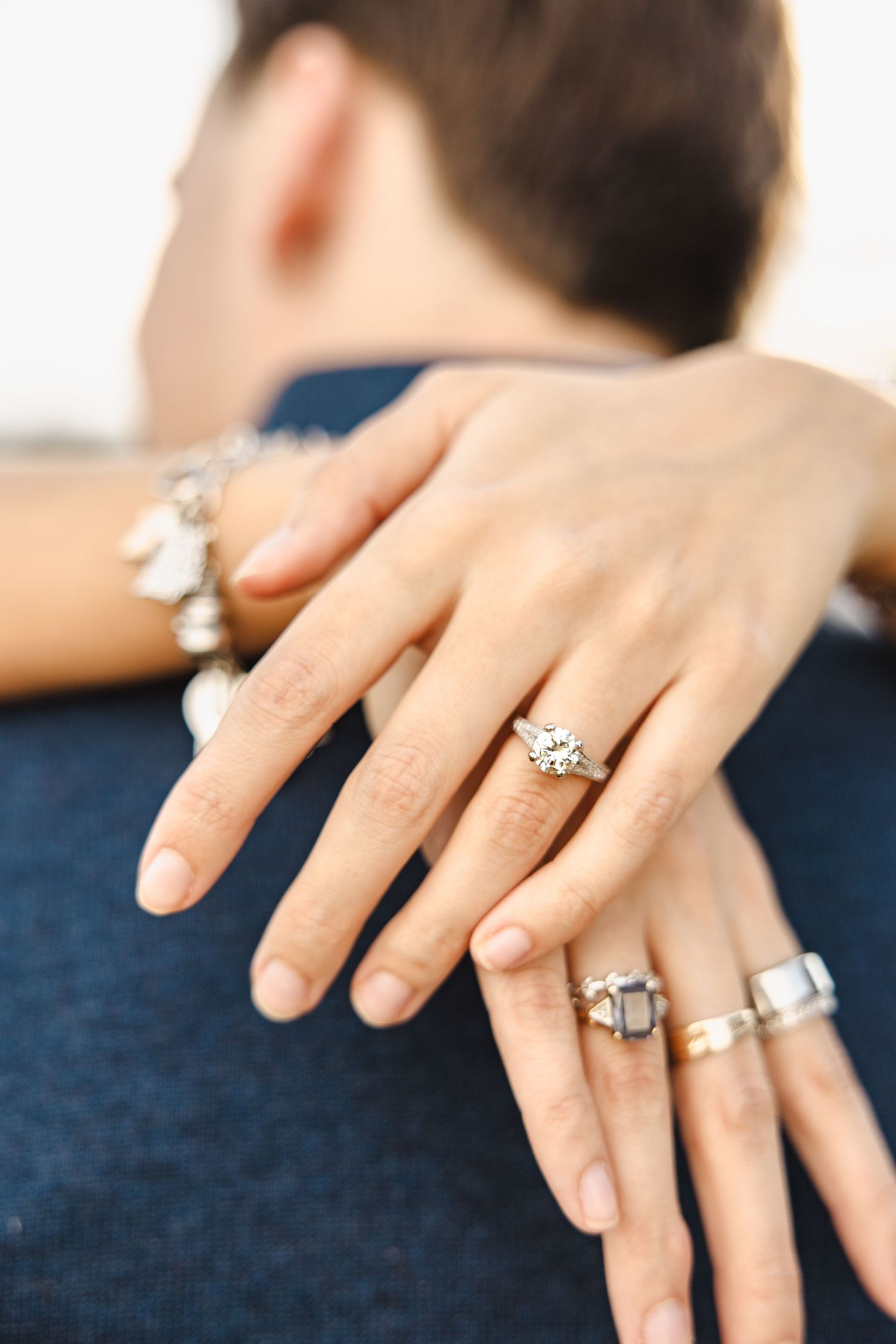 Close-up of womans hands showing diamond engagement ring and jewelry on mans shoulder.