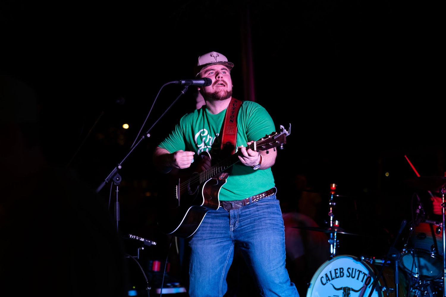 Smiling performer in green shirt plays guitar and sings at a live show with band visible behind him.