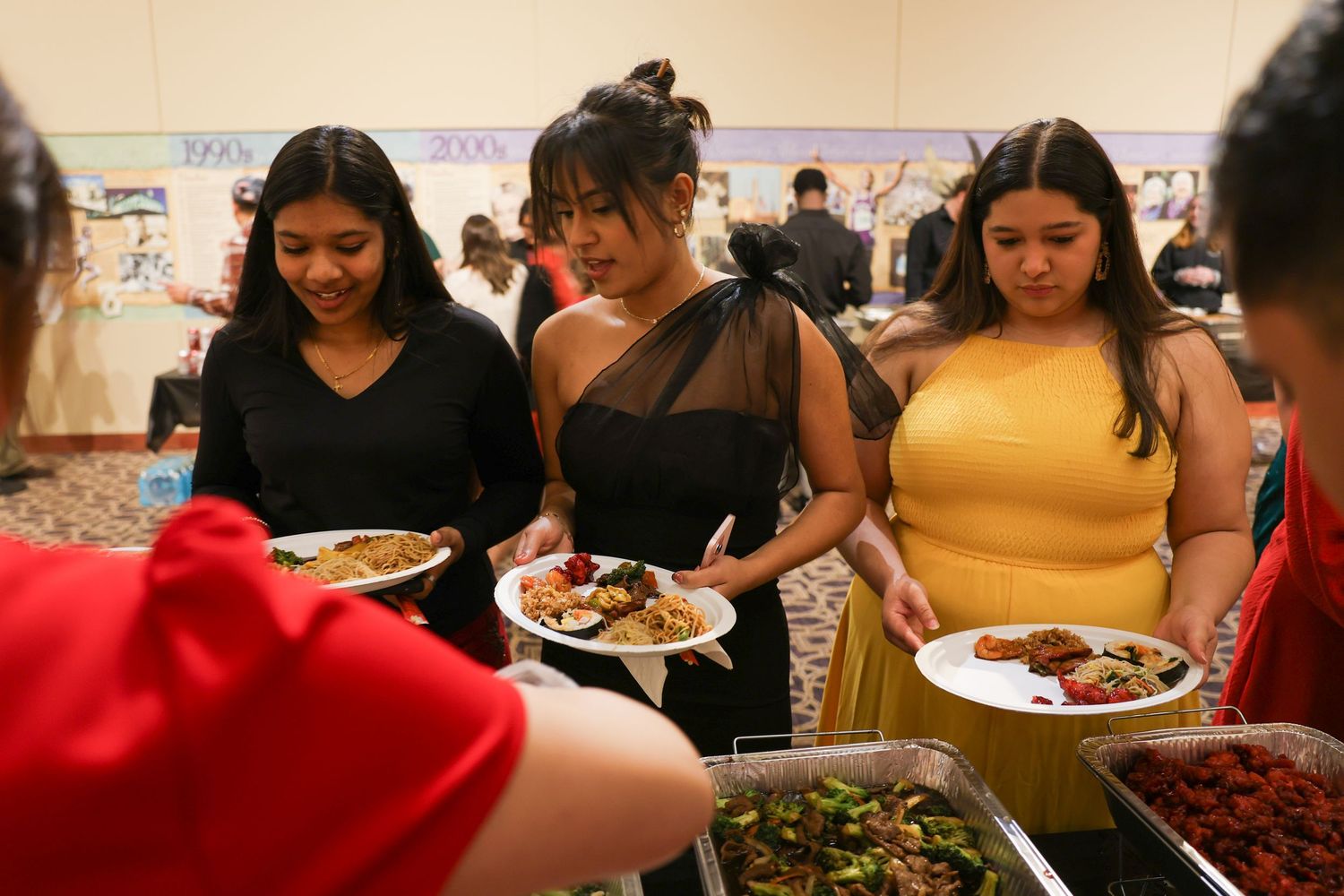 Three young women in elegant dresses serving themselves food at a festive buffet celebration.