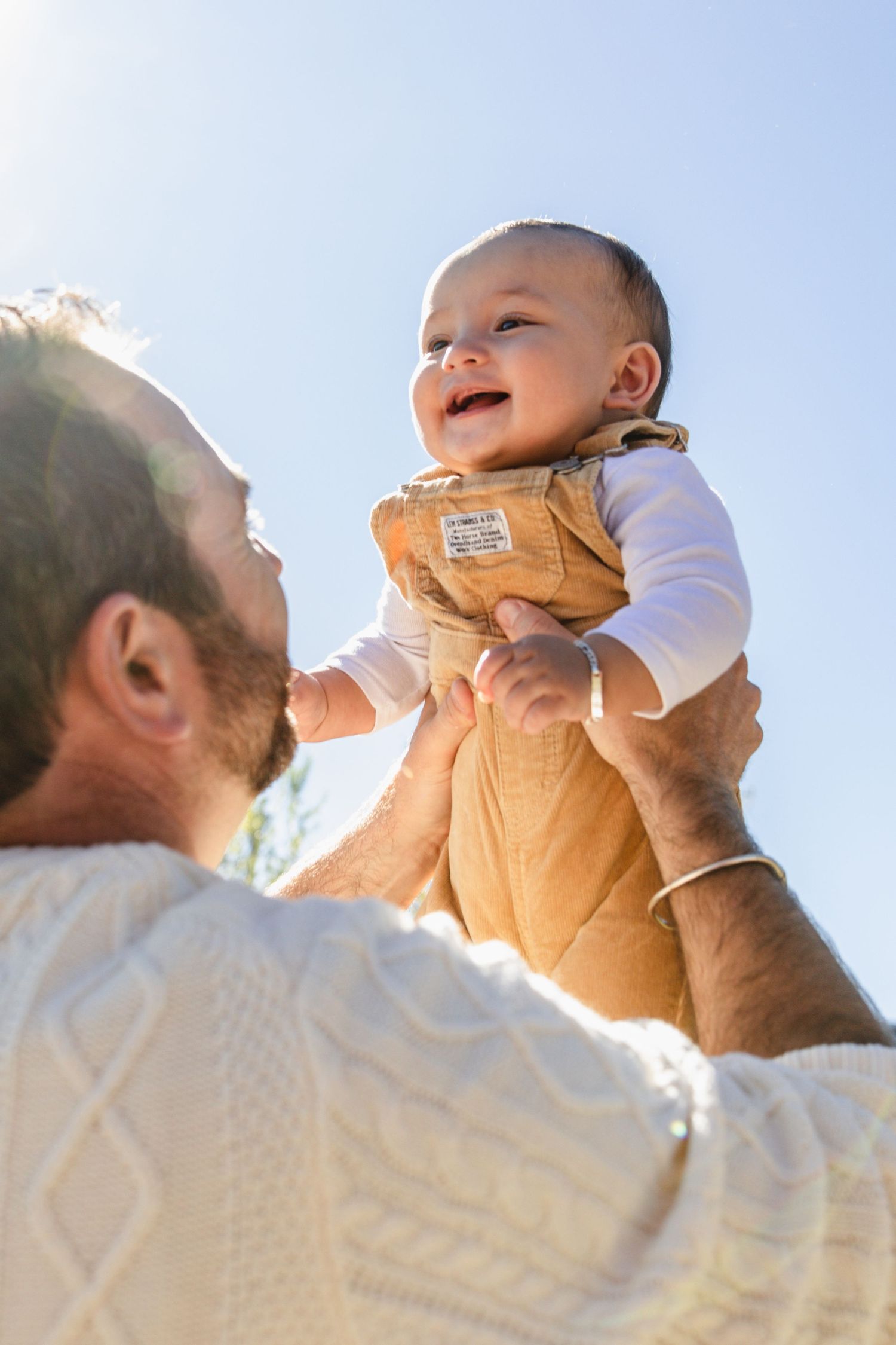 Happy father lifting smiling baby in tan vest against a bright blue sky outdoors.