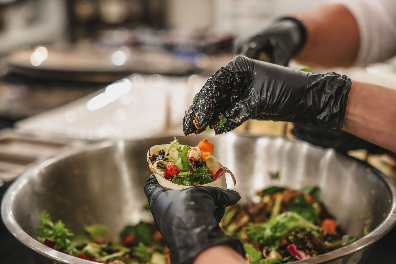 Chef in black gloves assembling a fresh taco with colorful vegetables over a large mixing bowl.