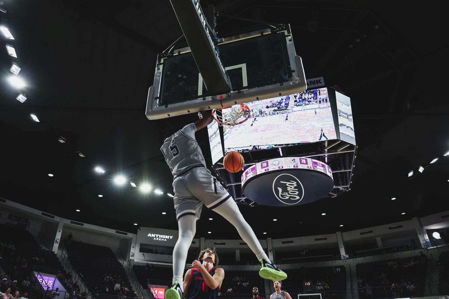 Basketball player dunks over the rim with a large scoreboard screen visible in a packed indoor arena.