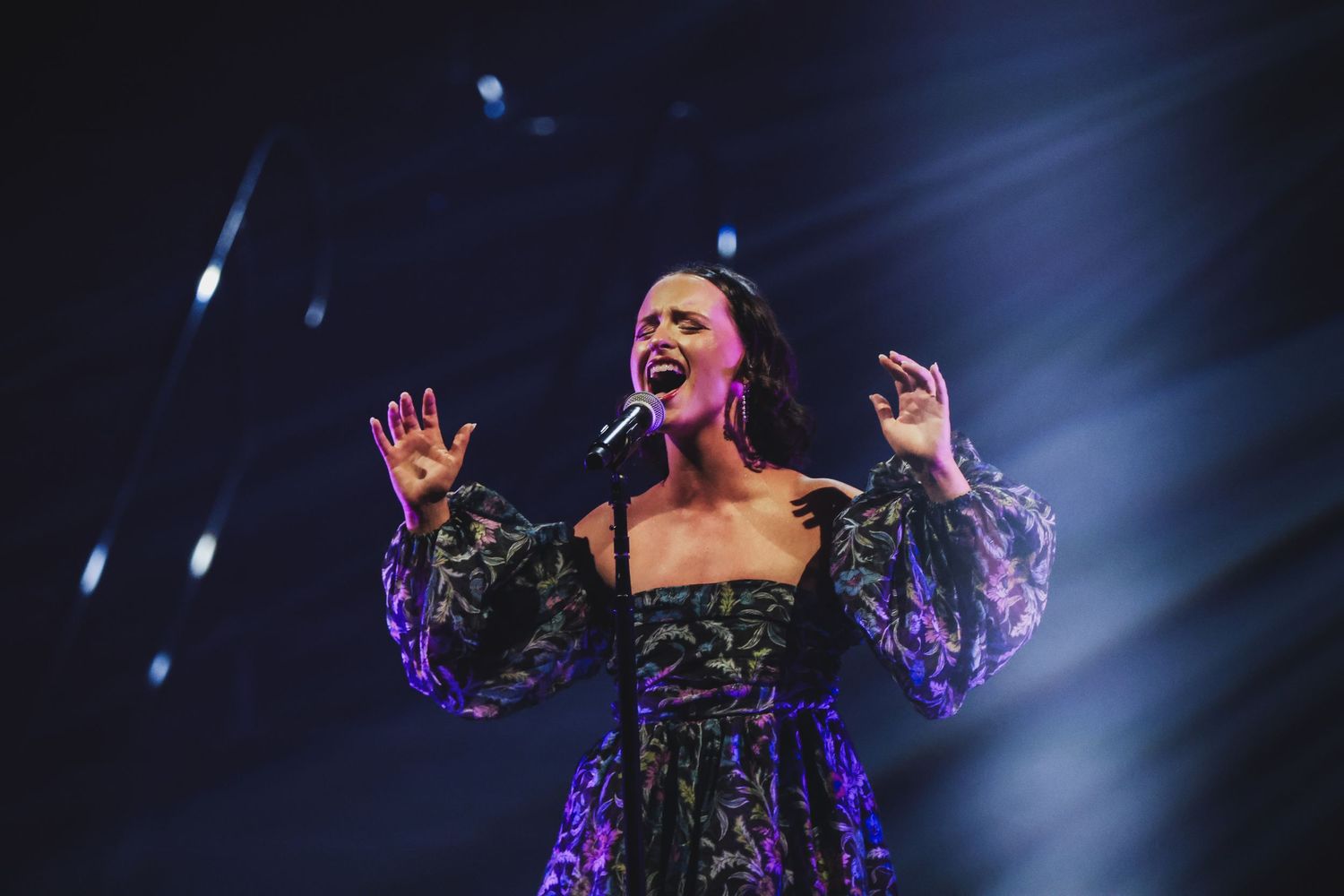 Female vocalist in black floral dress singing passionately on stage under purple spotlights.