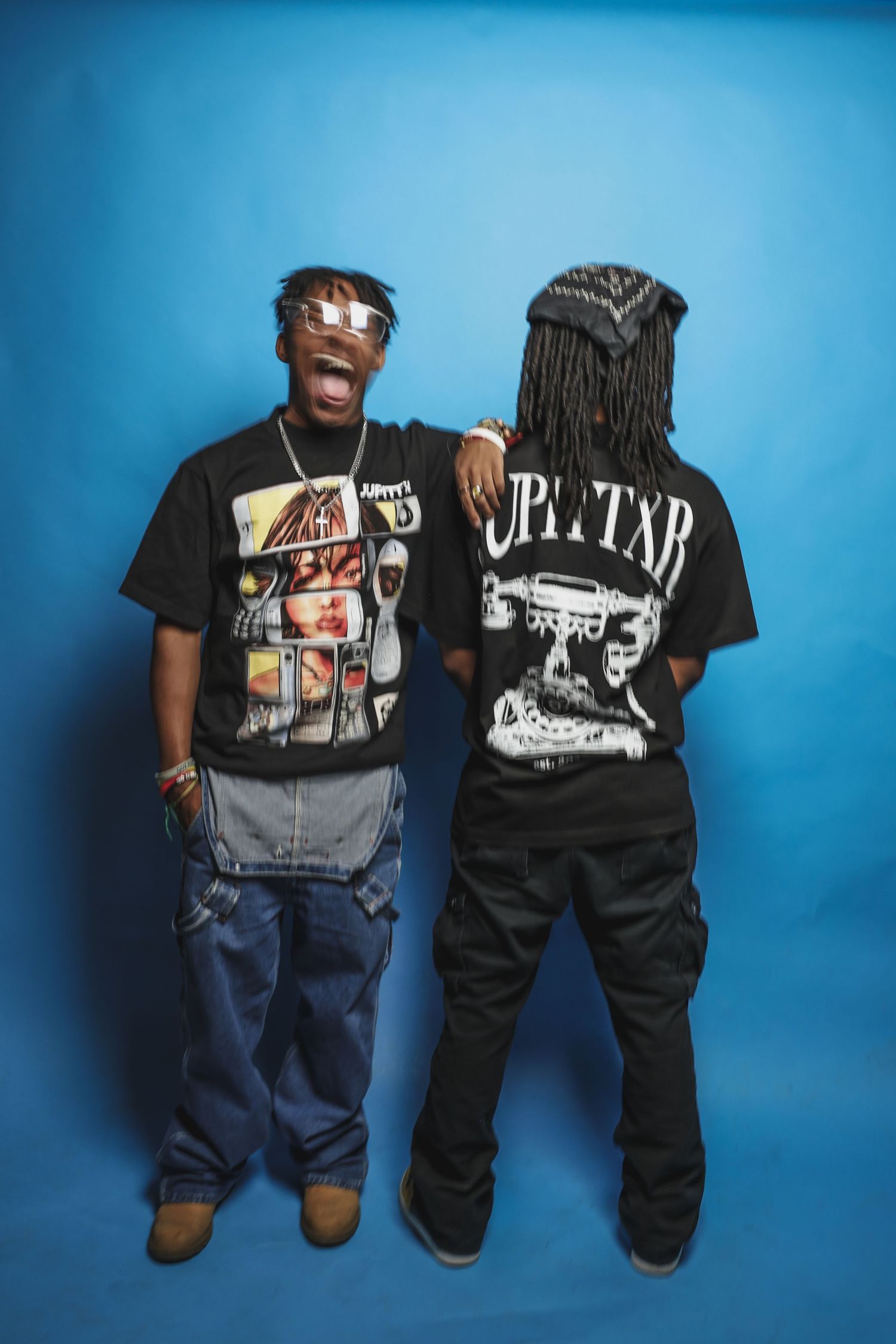 Two men in graphic black tees pose back-to-back against a bright blue studio backdrop.