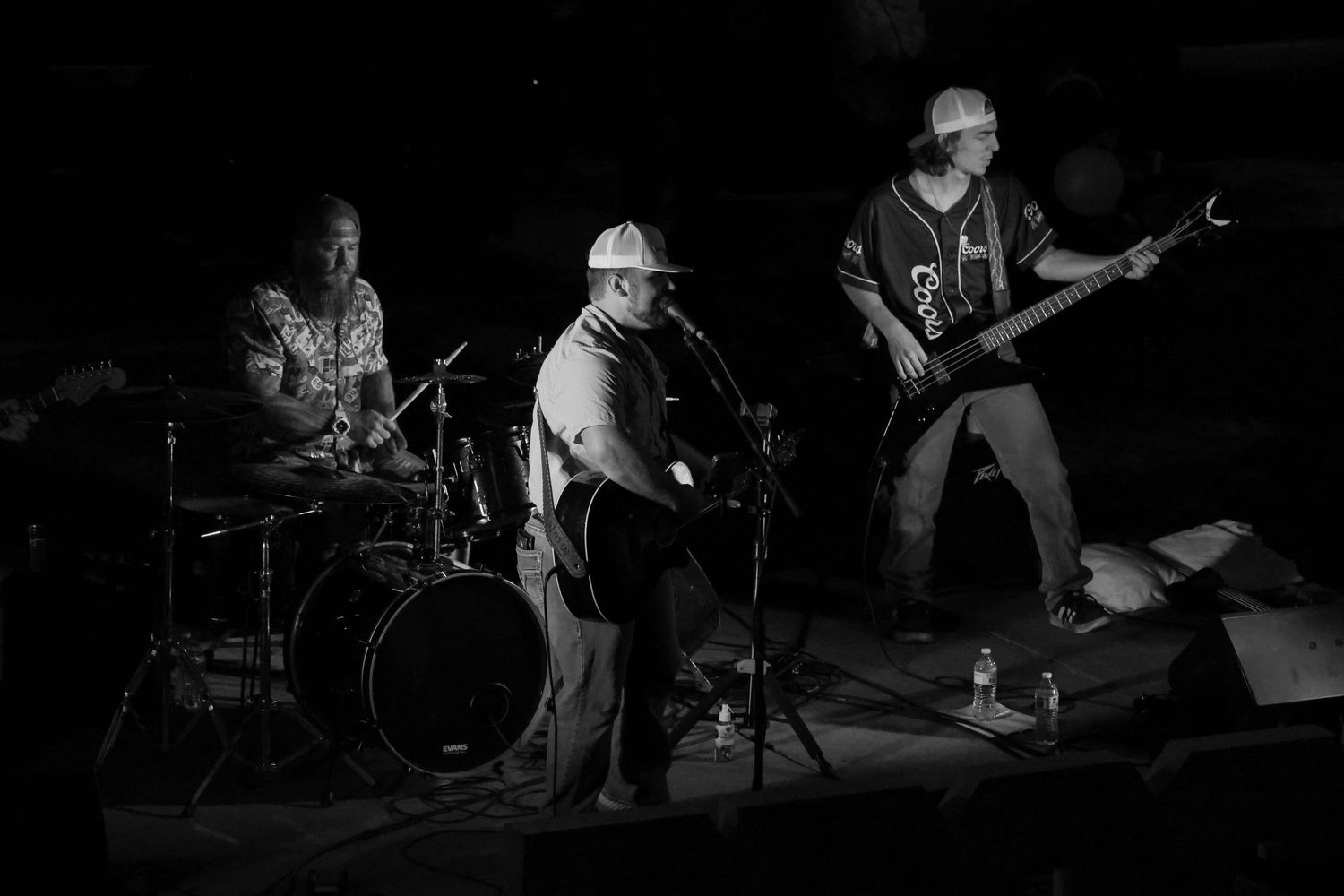 Black and white photo of a three-piece band performing on a dimly lit indoor stage.