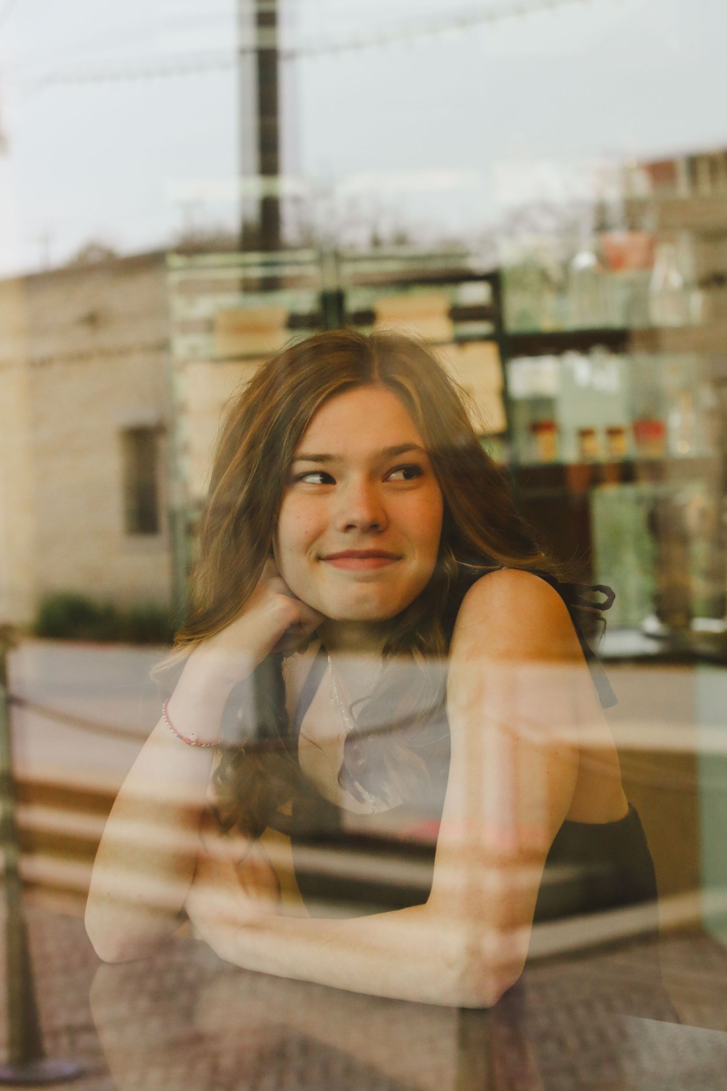Young woman smiling thoughtfully through a cafe window, resting her chin on her hand in a candid portrait.