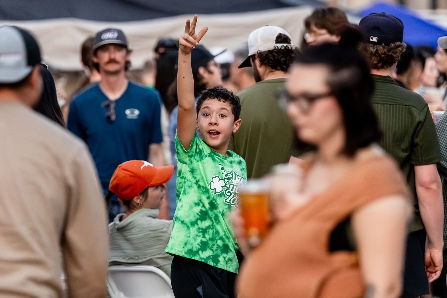 Excited young boy in green tie-dye shirt waves at a festive St. Patricks Day outdoor festival.