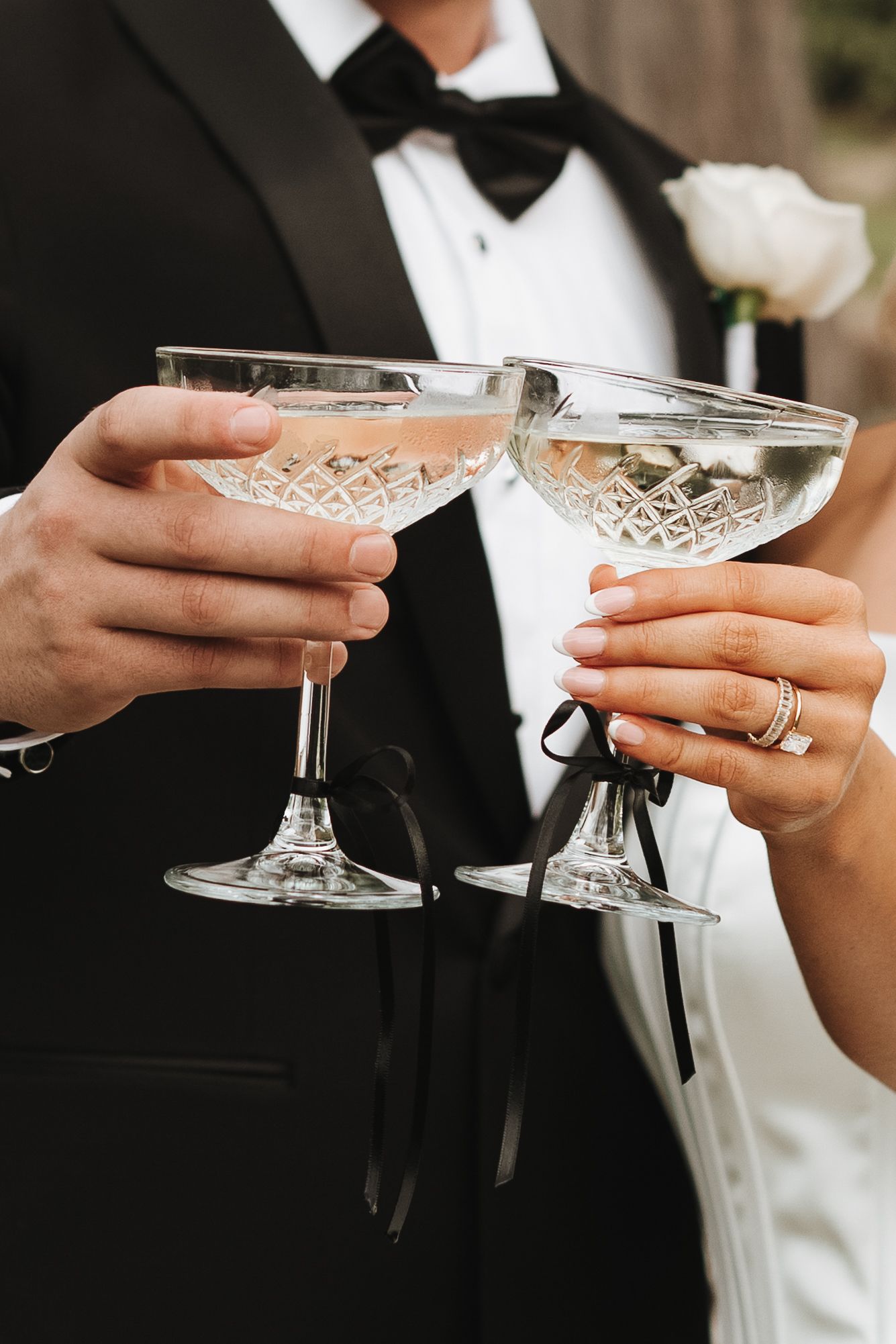 Bride and groom holding crystal champagne coupe glasses with black ribbon accents at a formal wedding celebration.