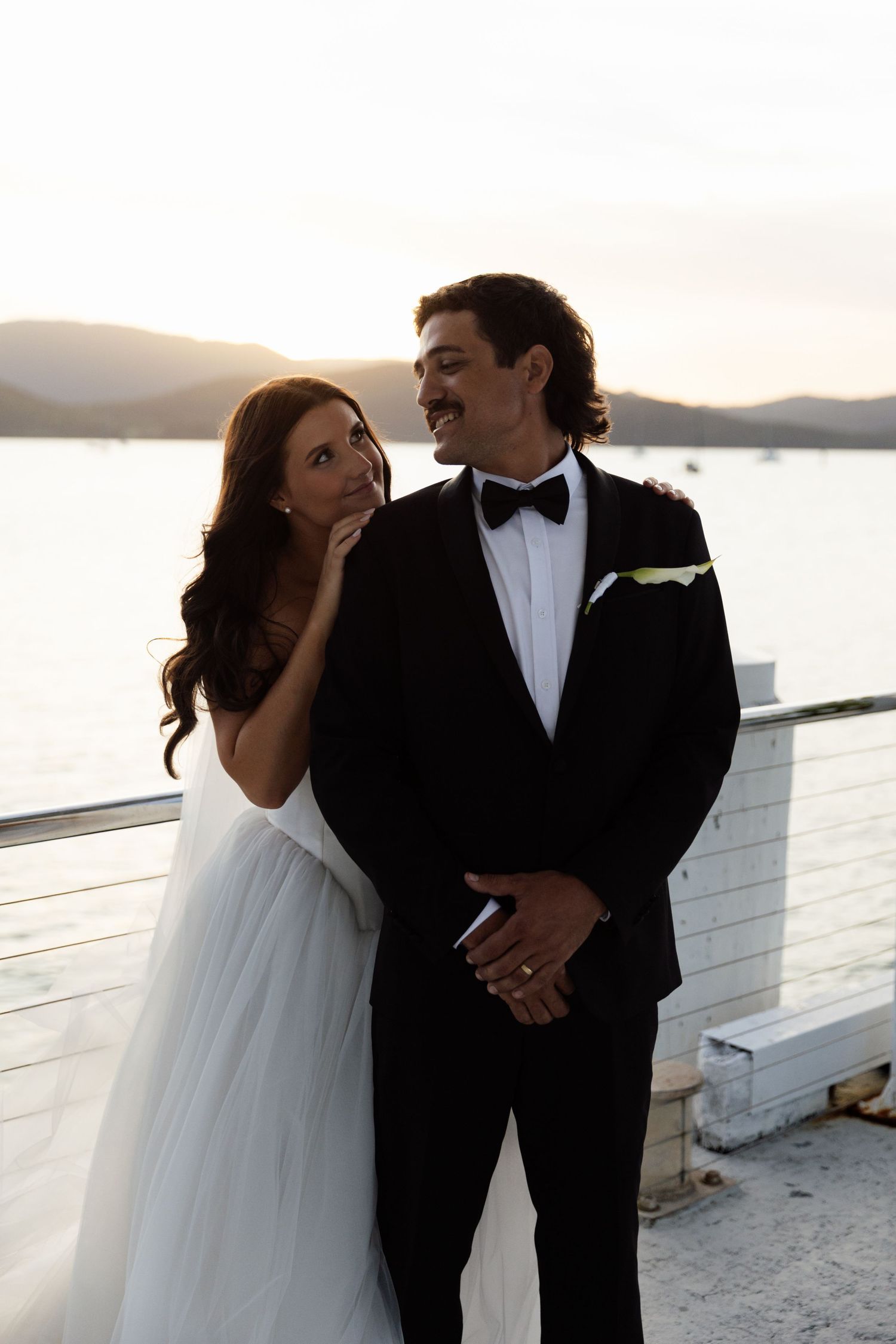 Bride and groom in wedding attire pose on waterfront deck at sunset with mountains in background.