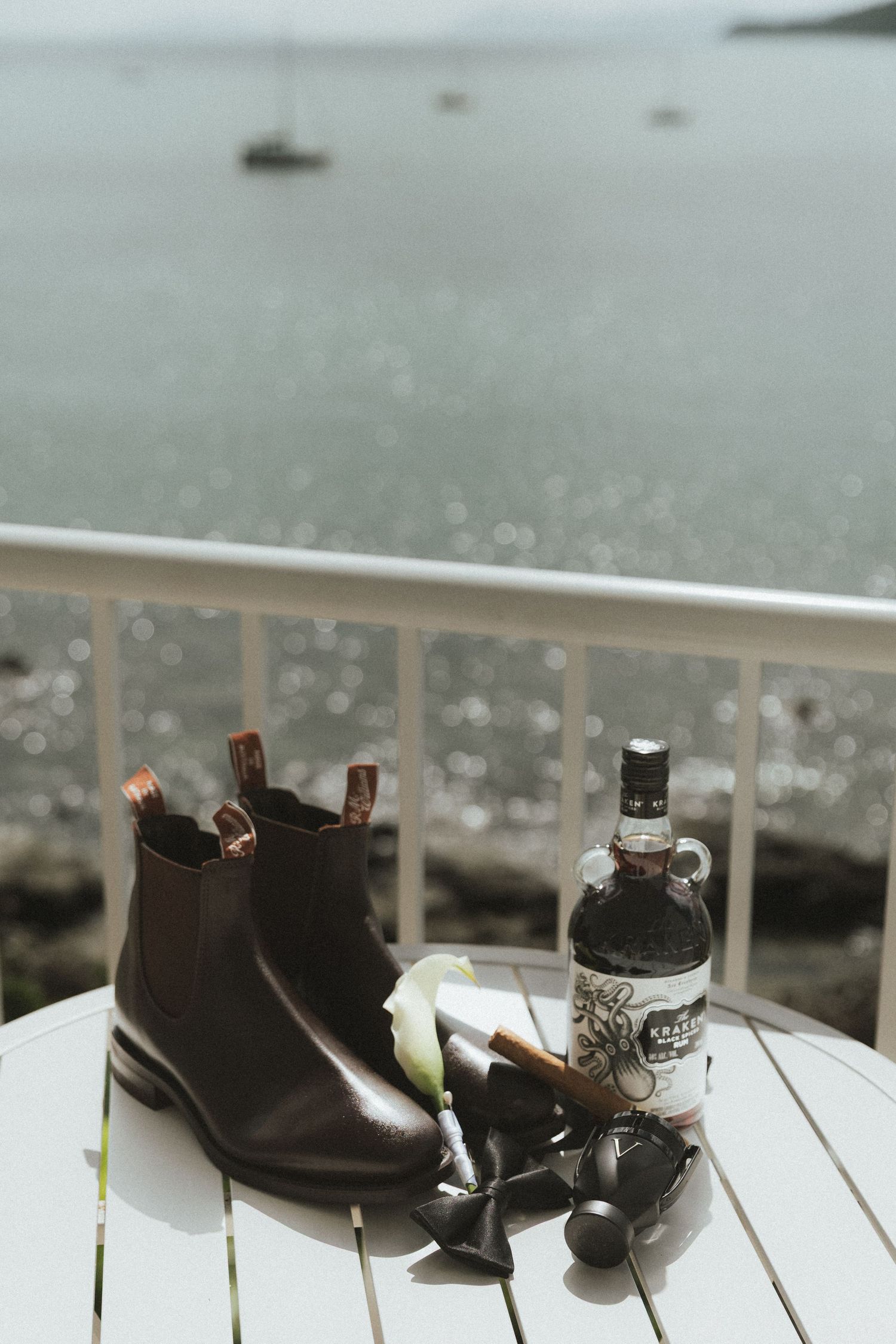 Grooms Chelsea boots, whiskey bottle, and boutonniere on white table overlooking ocean.