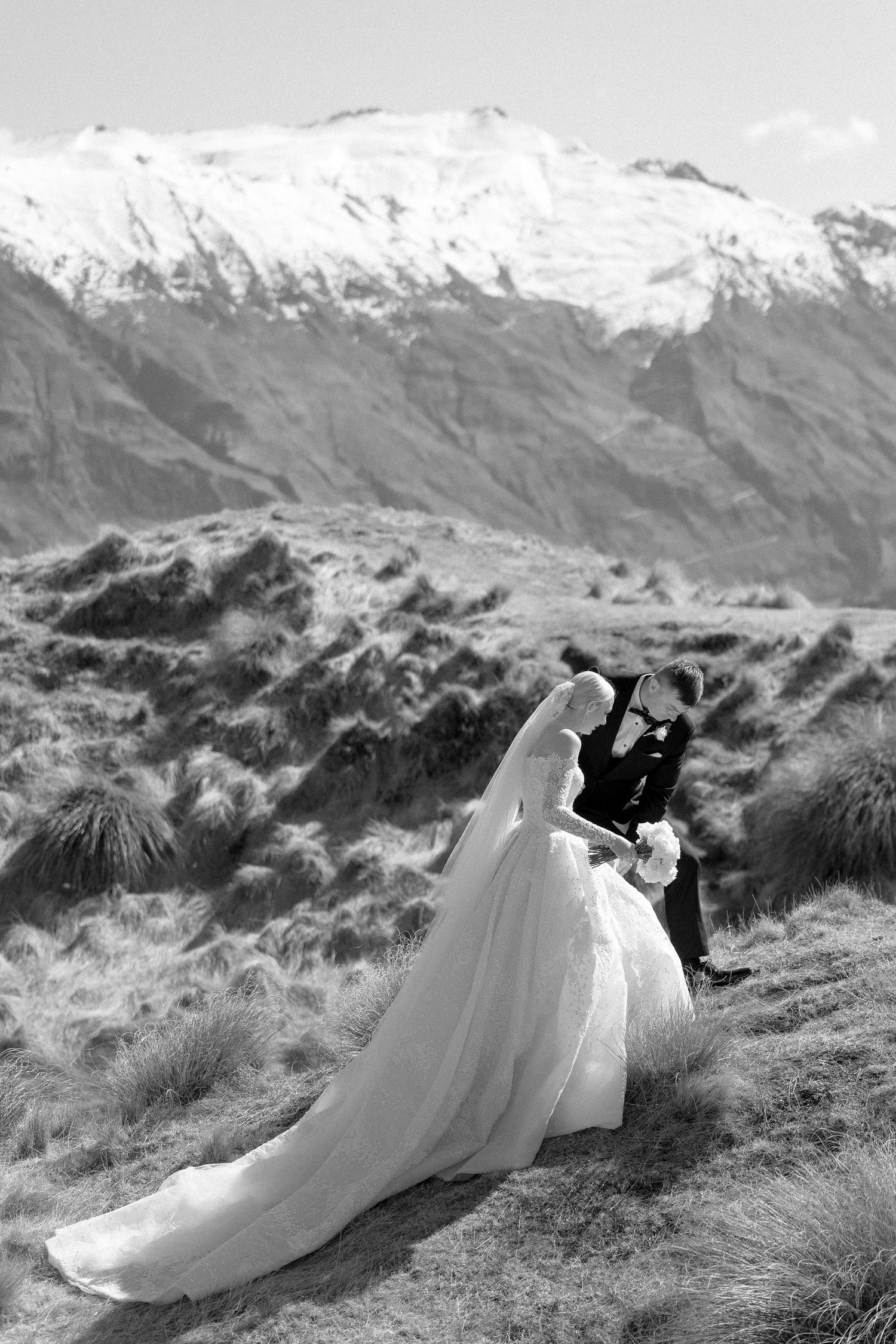 Black and white wedding photo of bride and groom kissing on a mountain with snowy peaks backdrop.