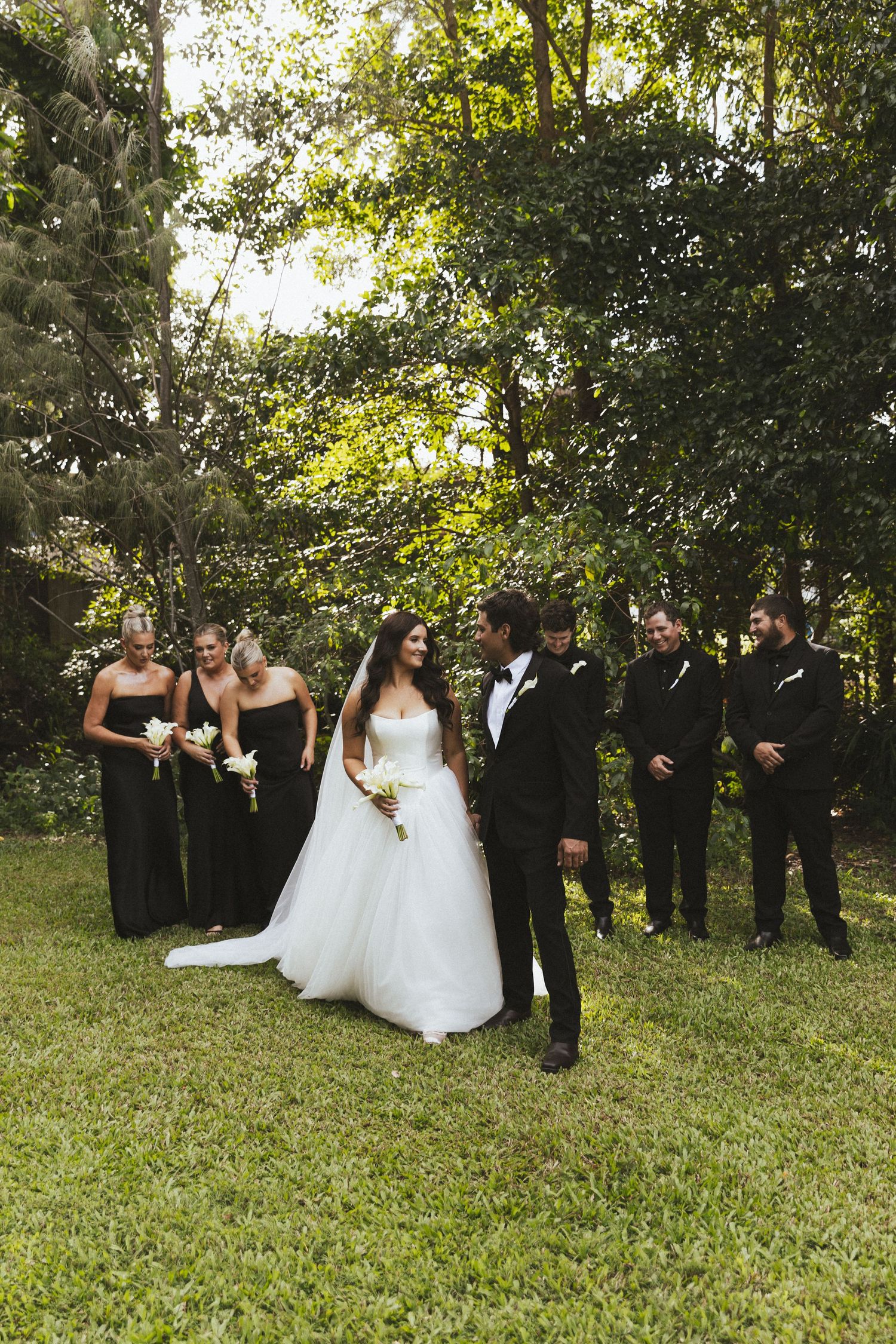 Wedding party in black attire stands on green lawn with lush trees as bride and groom share a romantic moment.