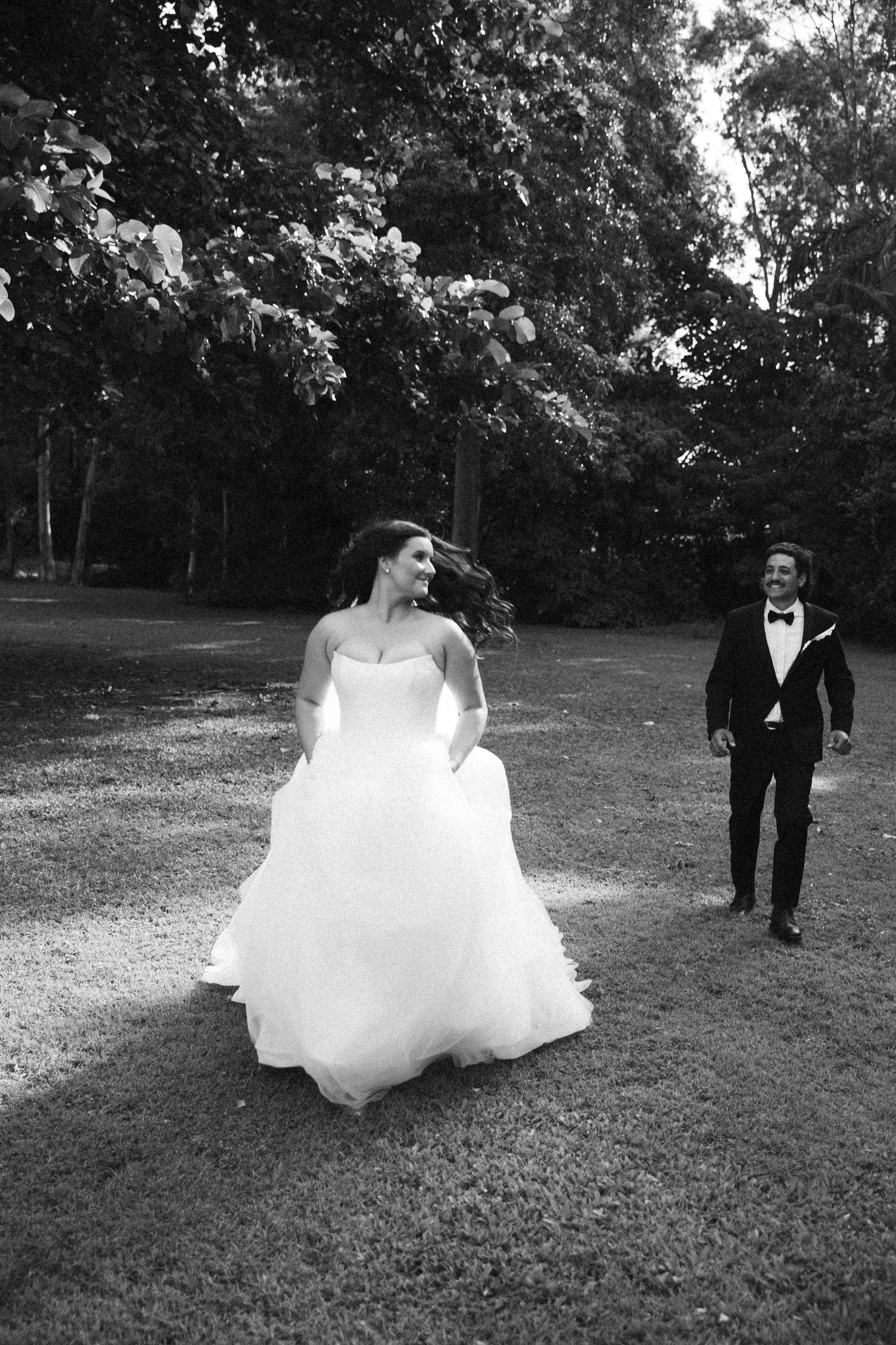 Black and white photo of joyful bride lifting wedding gown while groom follows her across a garden path.