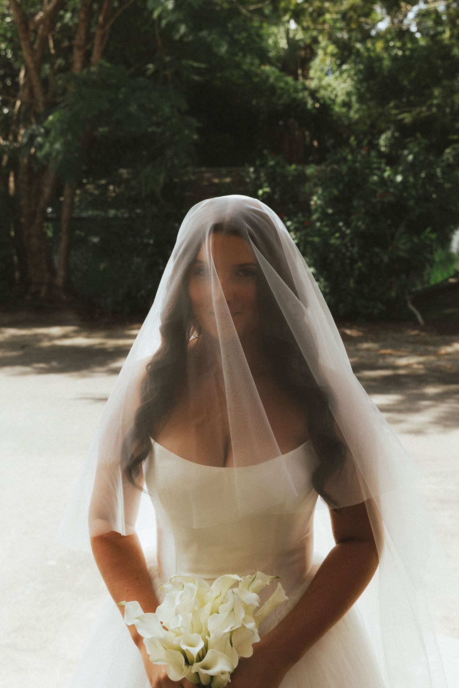 Elegant bride holding white hydrangea bouquet with veil draped over face standing in sunlit garden setting.
