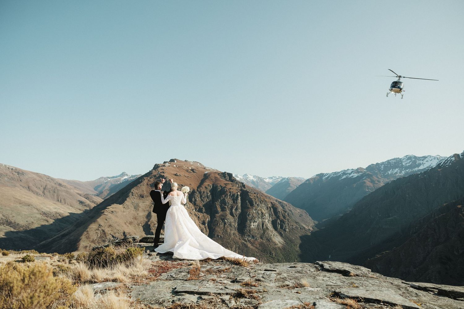 Bride and groom embrace on mountain peak with helicopter flying overhead against clear blue sky.
