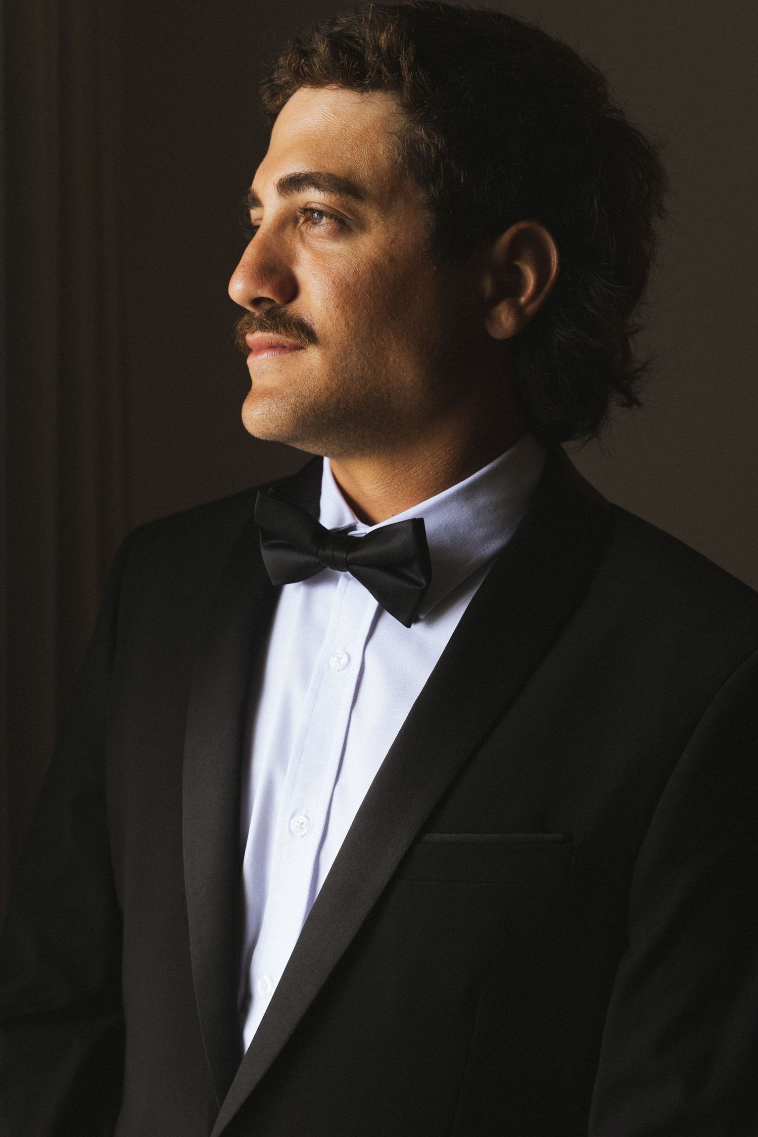 Dapper groom with mustache wearing a black tuxedo and bow tie gazes thoughtfully in dramatic studio lighting.