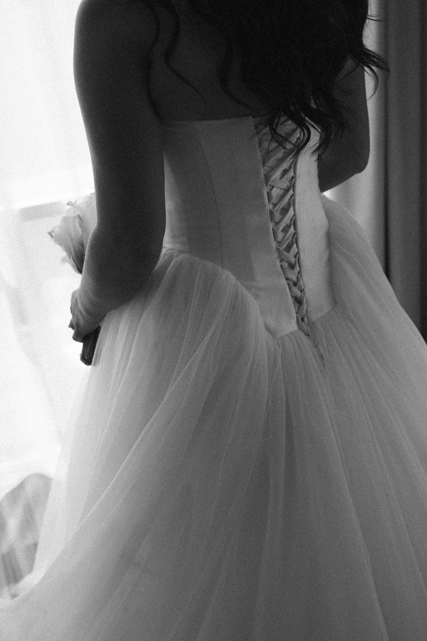 Black and white artistic back view of bride in corset-laced strapless tulle ball gown holding bouquet by window.