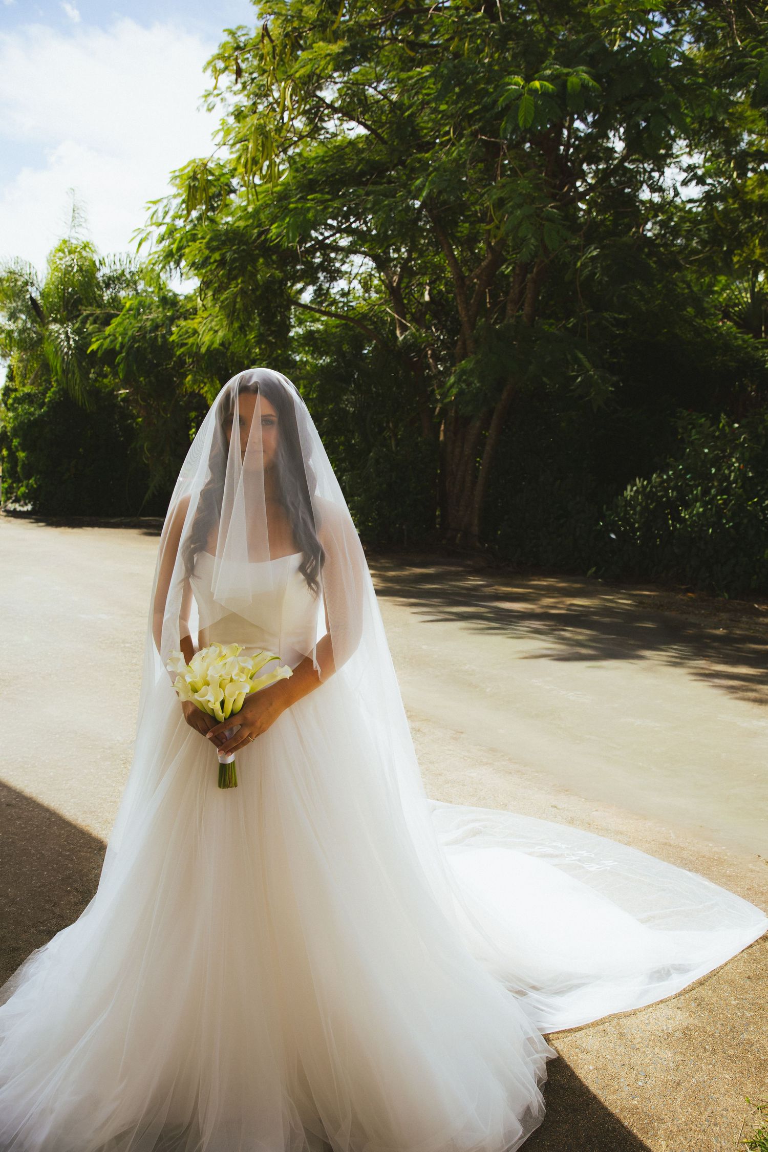 Bride in white tulle gown and veil holding yellow bouquet standing on sunlit path surrounded by lush green trees.