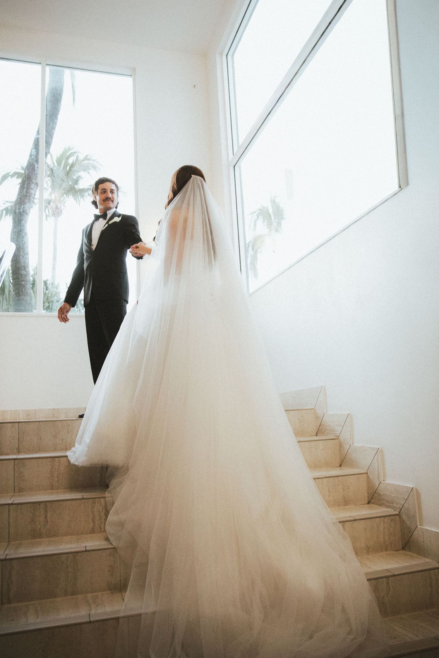 Bride in flowing tulle gown with long veil holds grooms hand while ascending marble staircase.