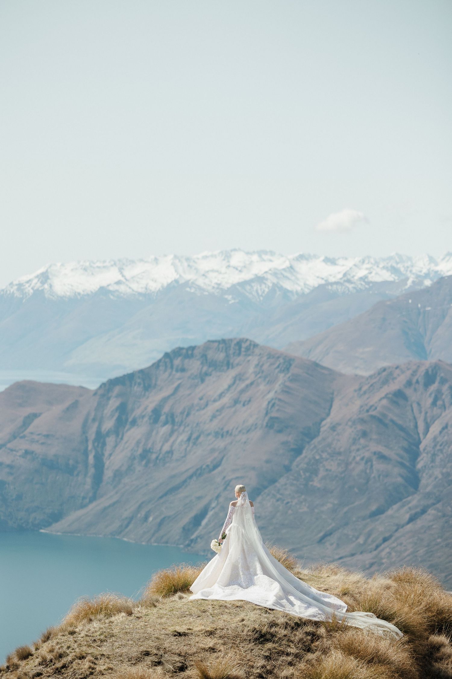 Bride in Steven Khalil gown with long train standing on mountain peak overlooking lake and mountains in Queenstown, New Zealand.