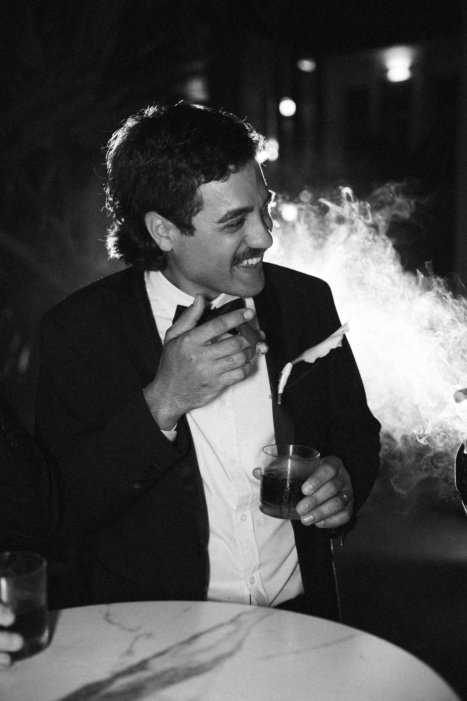 Black and white portrait of laughing groom in suit holding drink surrounded by smoke evening wedding reception.