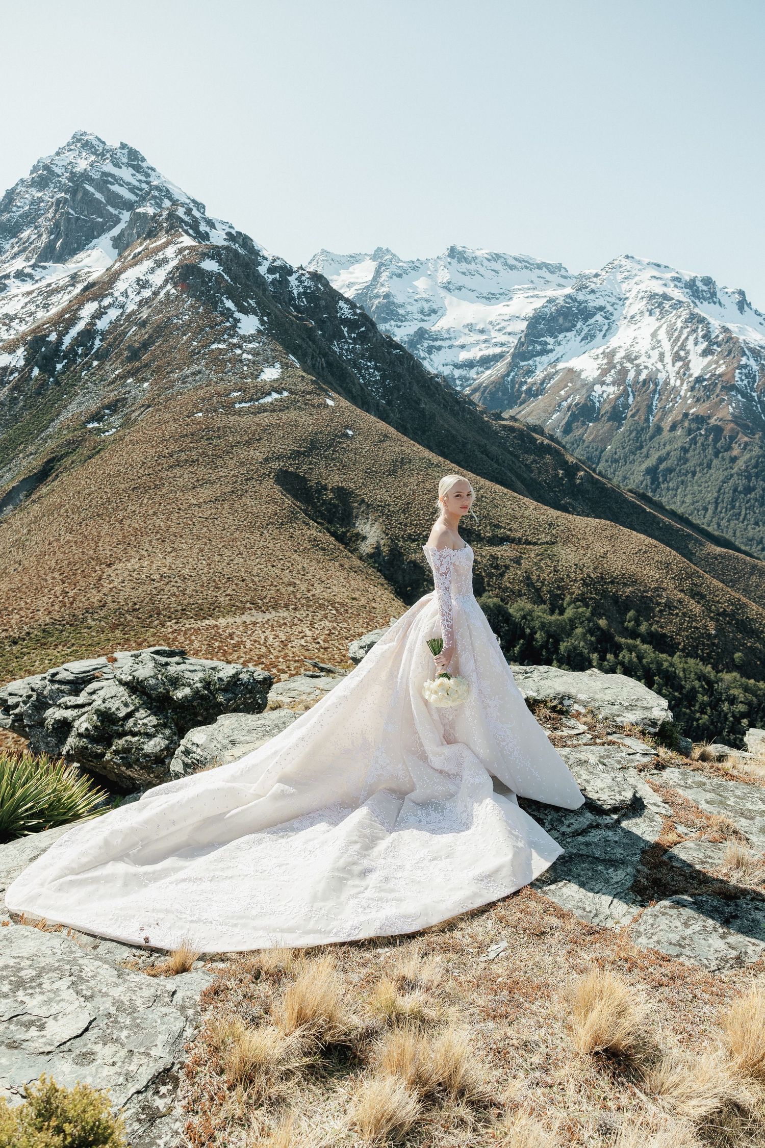 Bride in elegant french lace Steven Khalil gown with long train posing on rocky mountain summit in Queenstown, New Zealand.