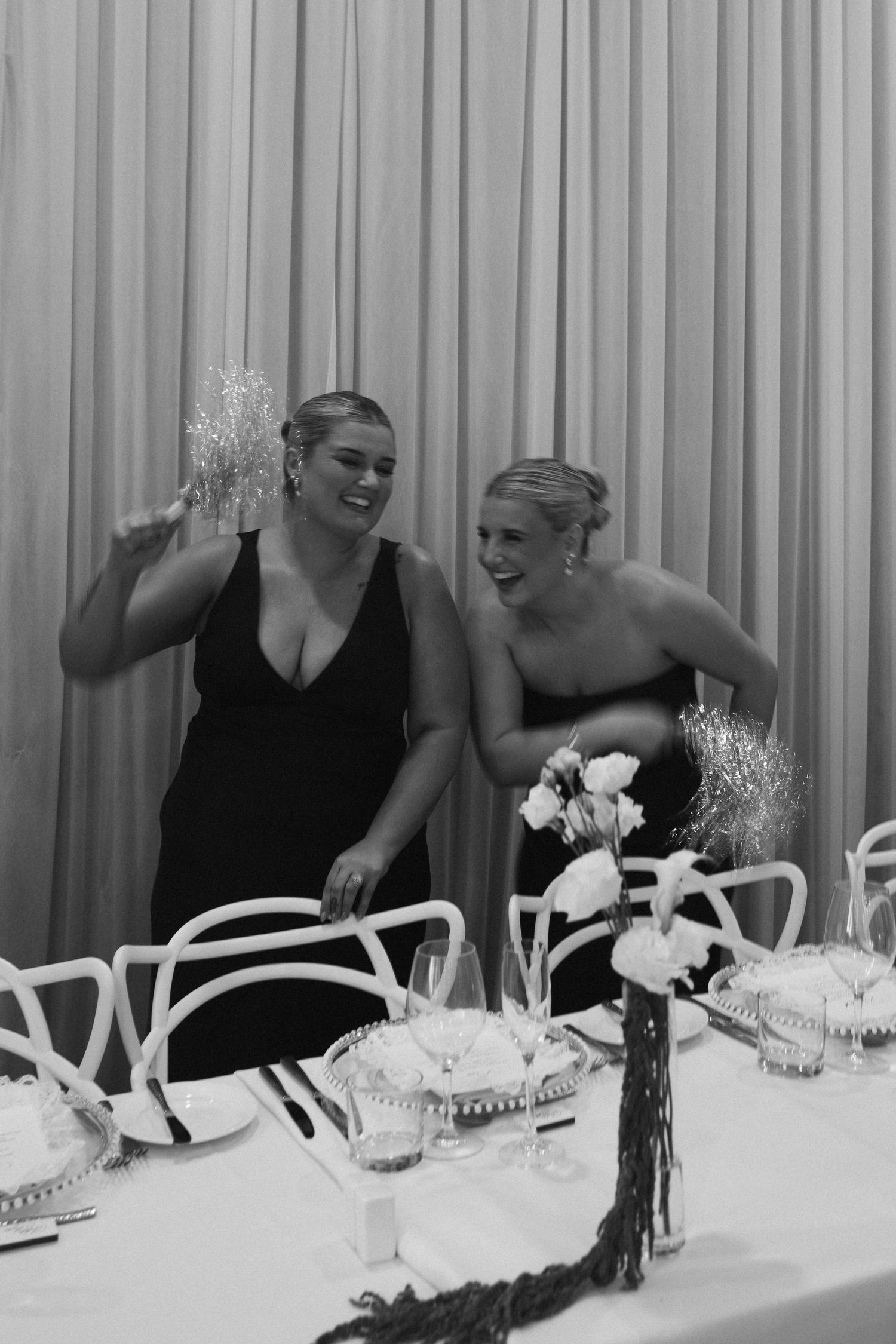 Two bridesmaids in black dresses laugh together at a wedding reception table in black and white photo.