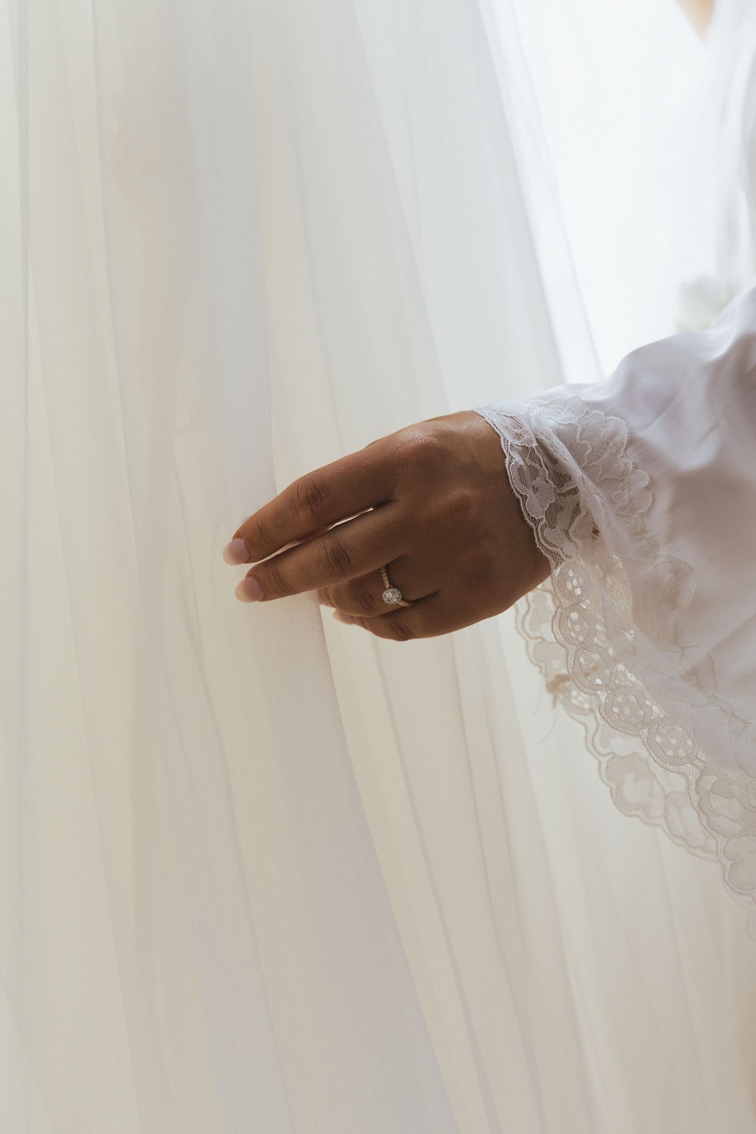 Bride wearing lace robe touches sheer wedding veil with diamond engagement ring on finger.