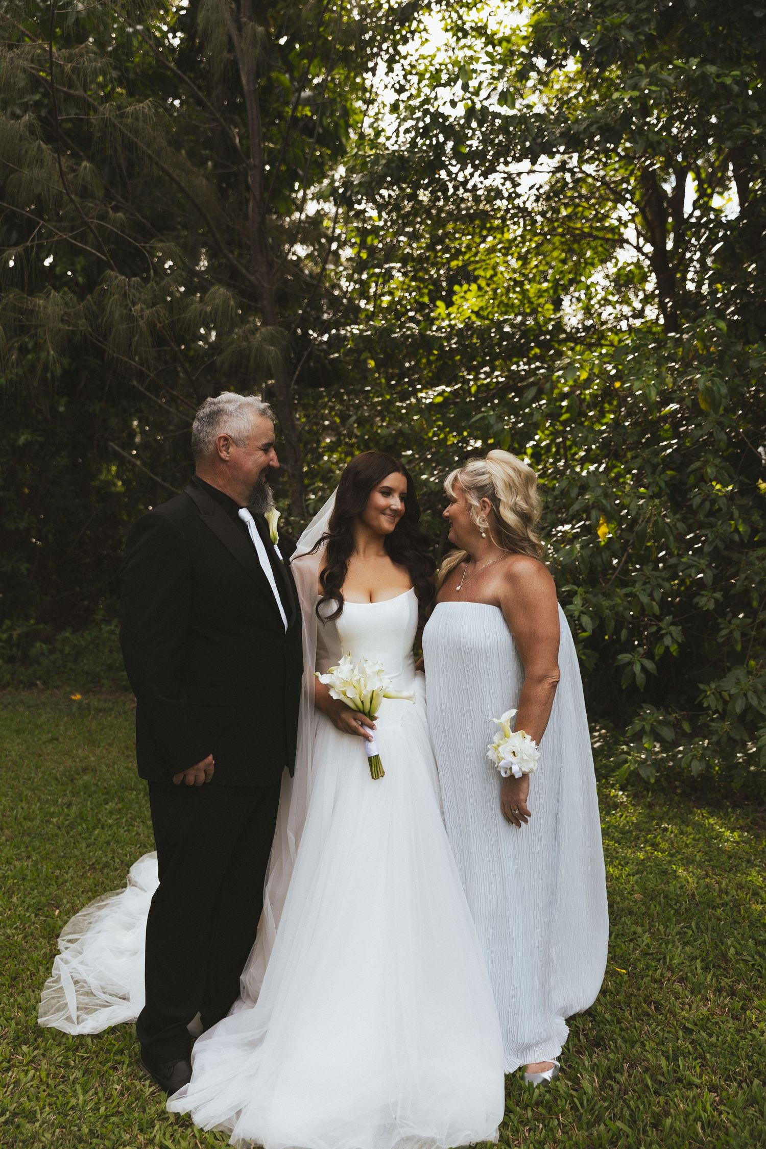 Bride in Aston Bridal gown holds bouquet while posing with parents in a lush green garden setting in Whitsundays, Queensland.