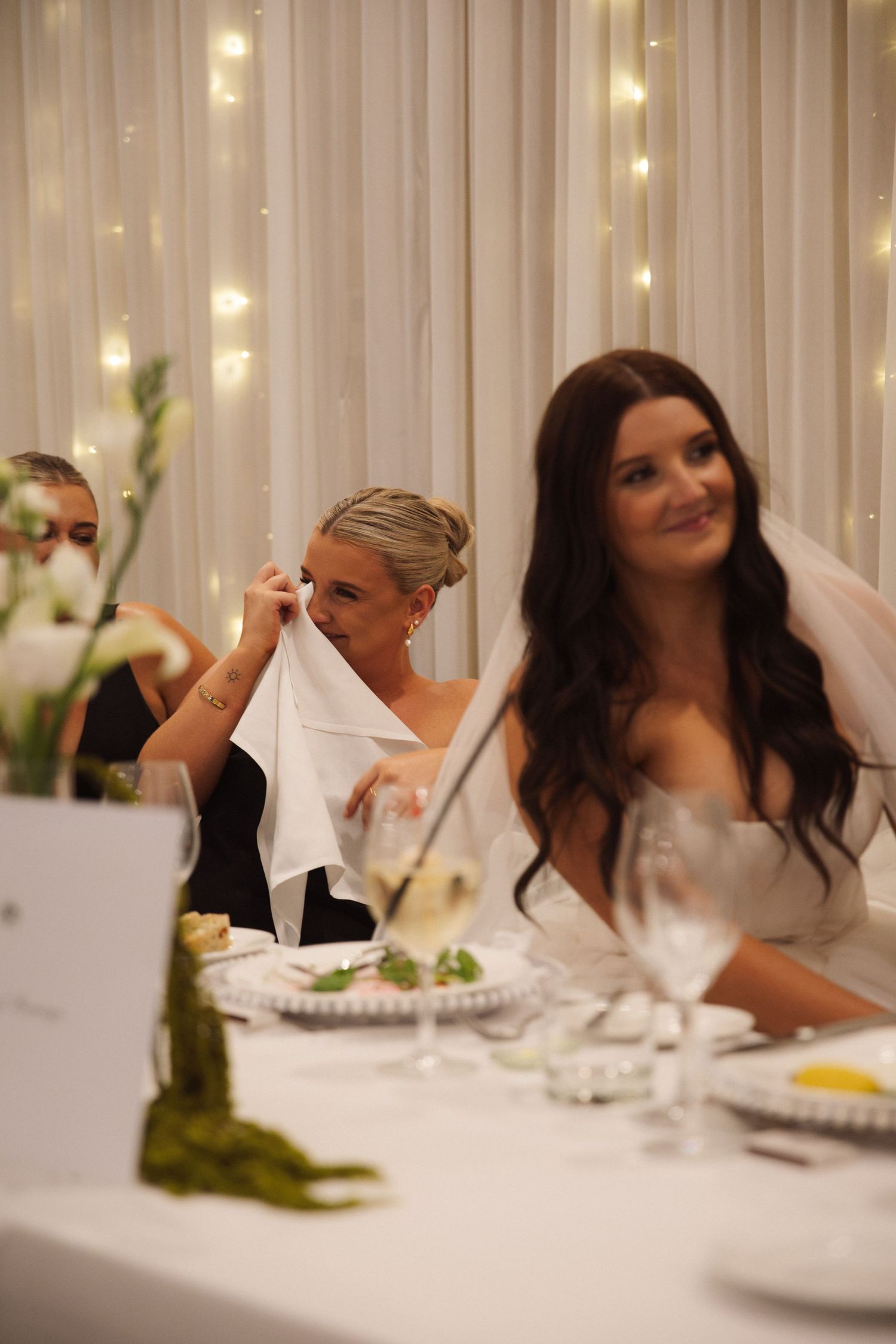 Emotional bridesmaid dabs tears with napkin beside smiling bride in white gown at wedding reception.