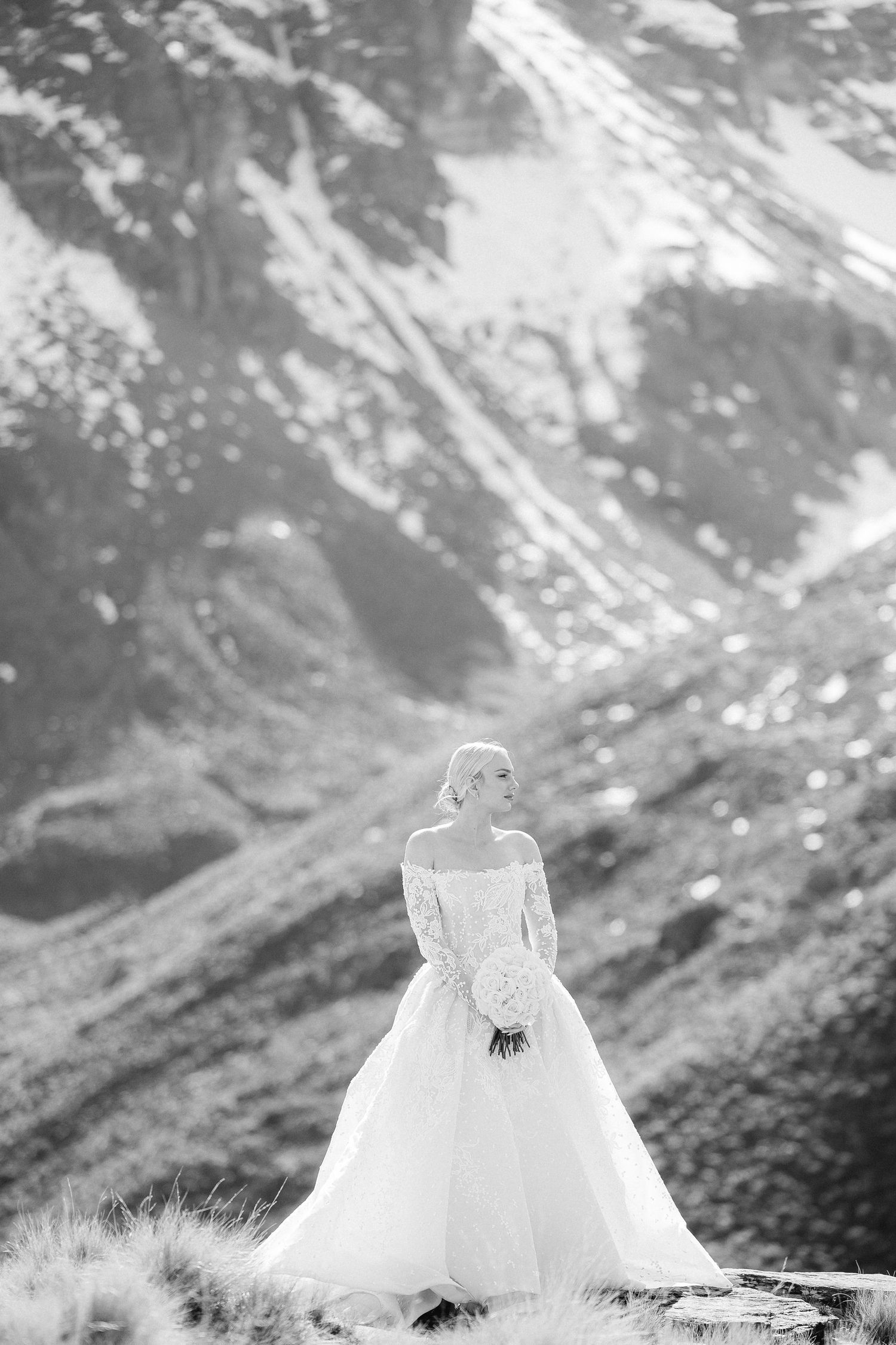 Black and white portrait of bride in lace Steven Khalil gown holding bouquet against dramatic mountain backdrop.