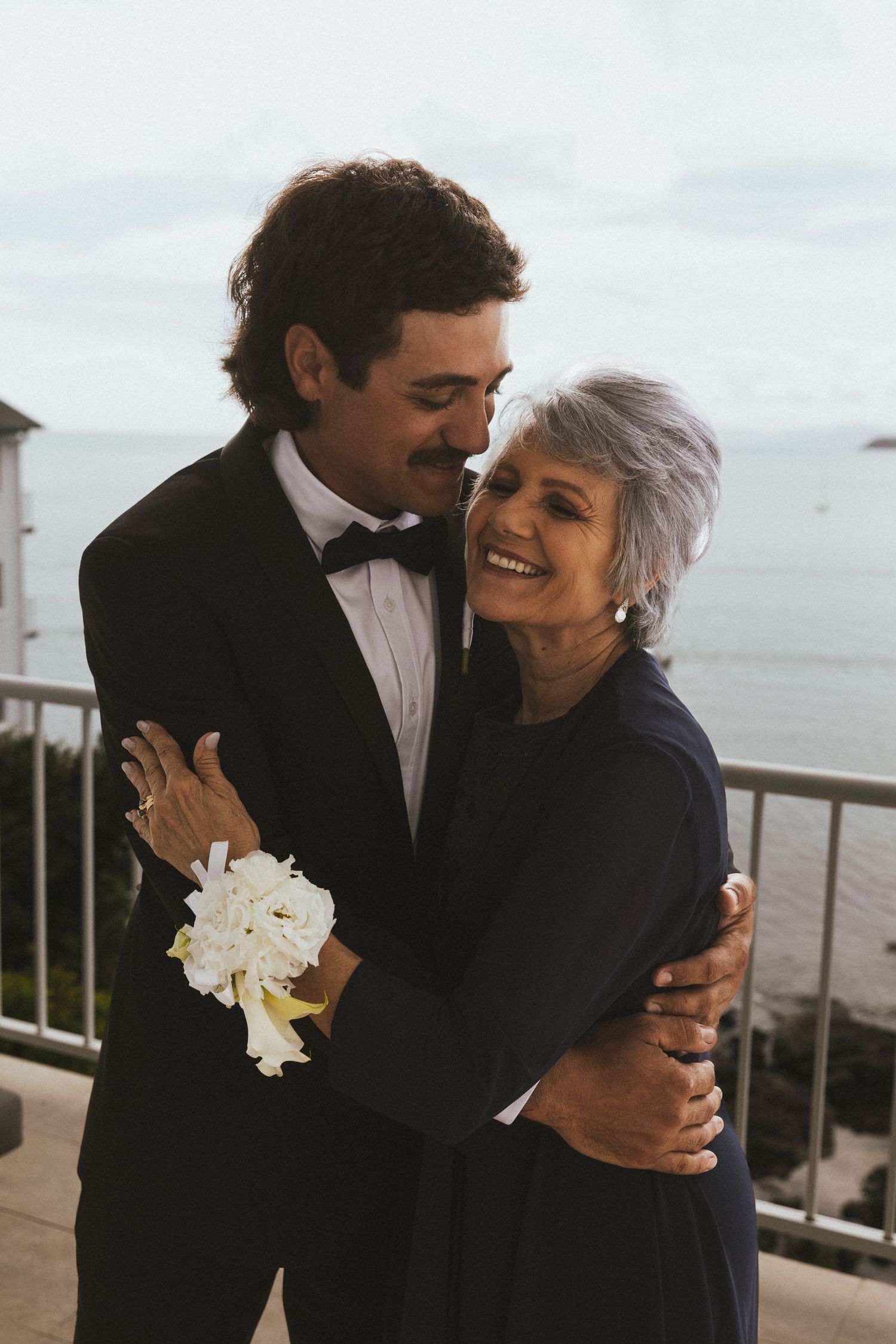 Groom in black tuxedo embracing laughing silver-haired mother wearing navy dress on balcony overlooking ocean.