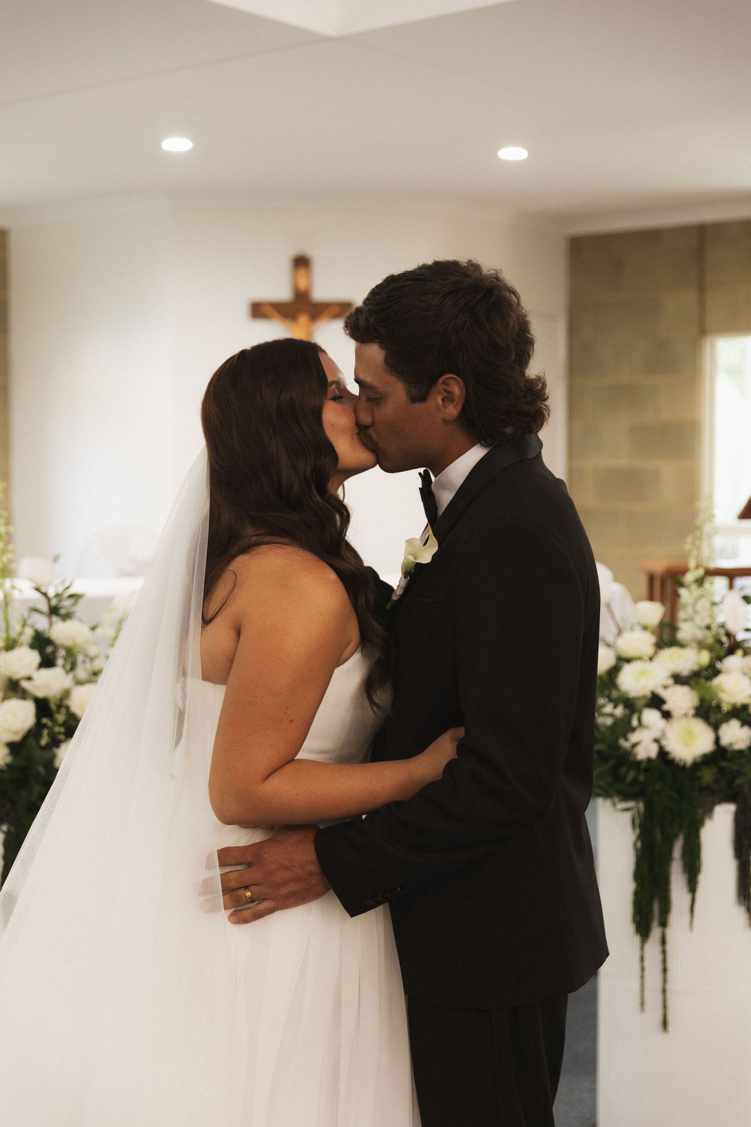 Bride and groom share first kiss at church altar decorated with white floral arrangements.